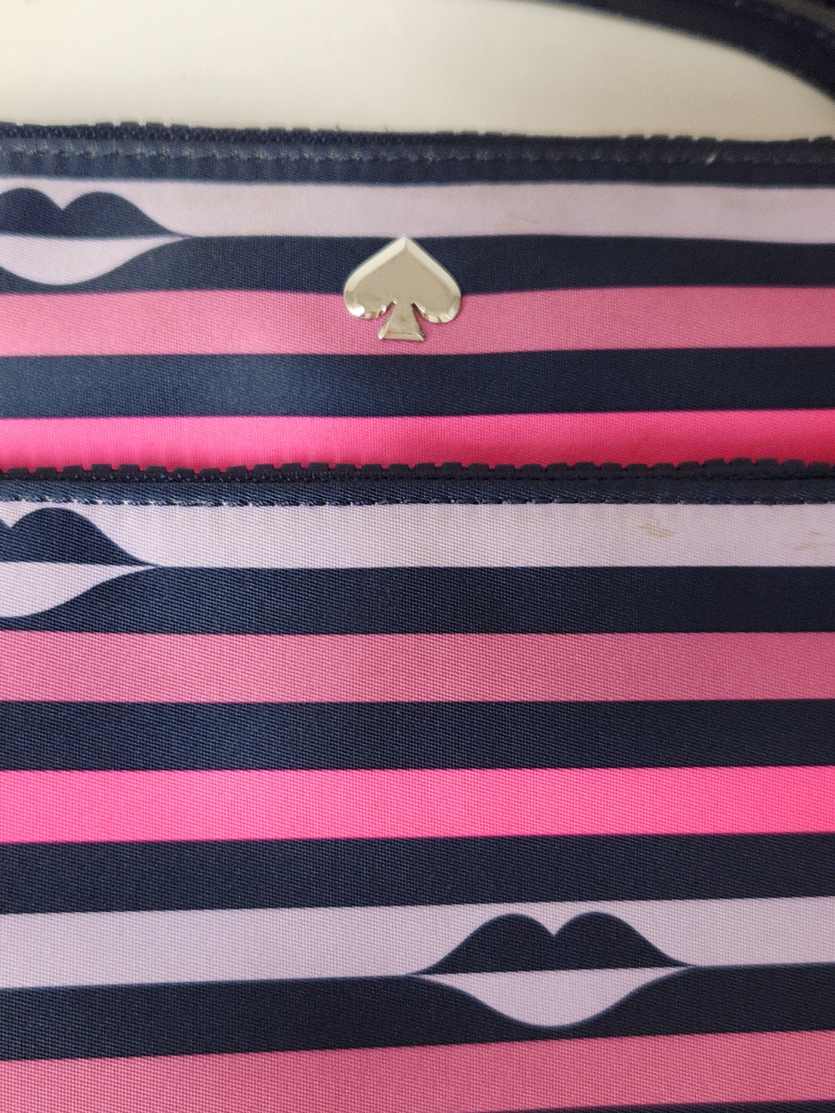 Close-up of a striped fabric accessory with navy, pink, and white horizontal bands, a small metallic spade-shaped logo at the top center, and lip motifs printed near the bottom.