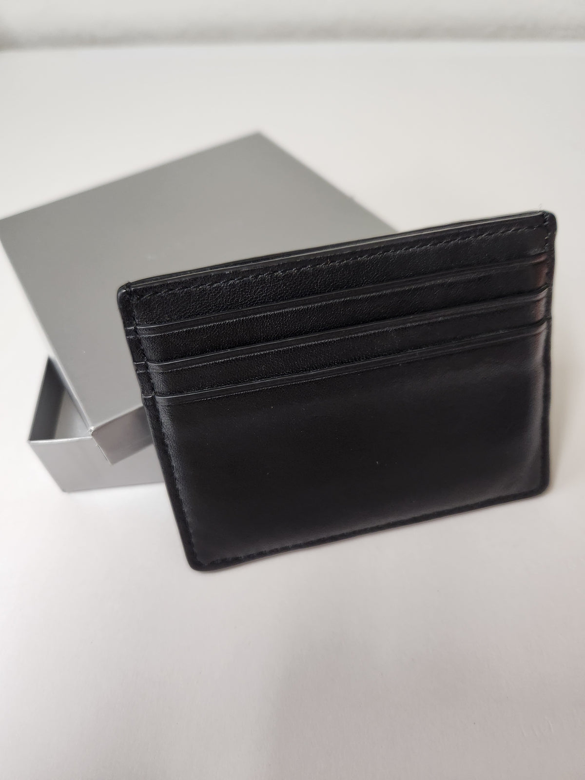 A black leather cardholder wallet standing upright next to its silver-gray presentation box on a light gray surface.