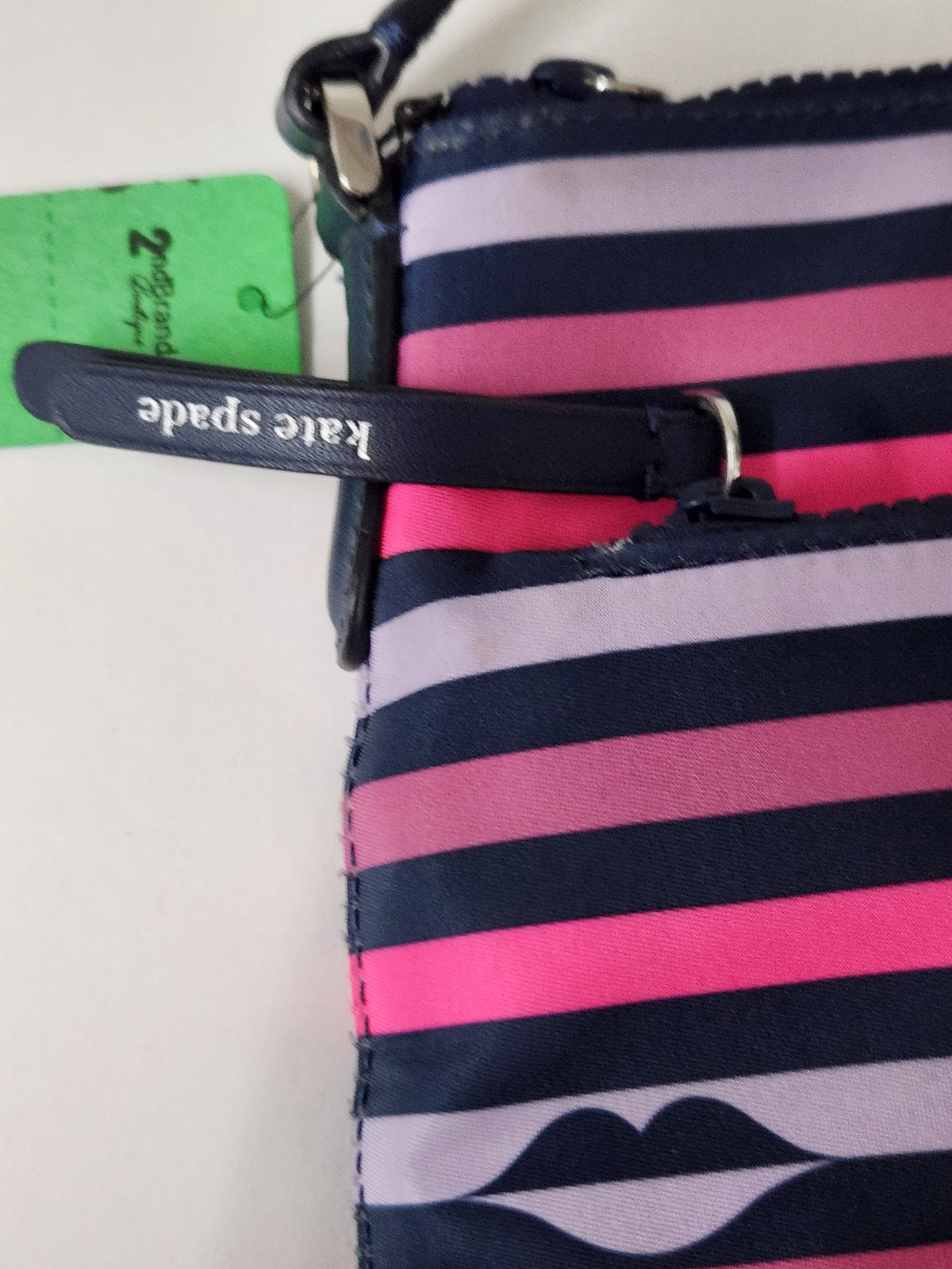 A close-up of a striped Kate Spade zip pouch or wallet in navy, pink, and light lavender, with a green tag and a black zipper pull labeled 'kate spade'.