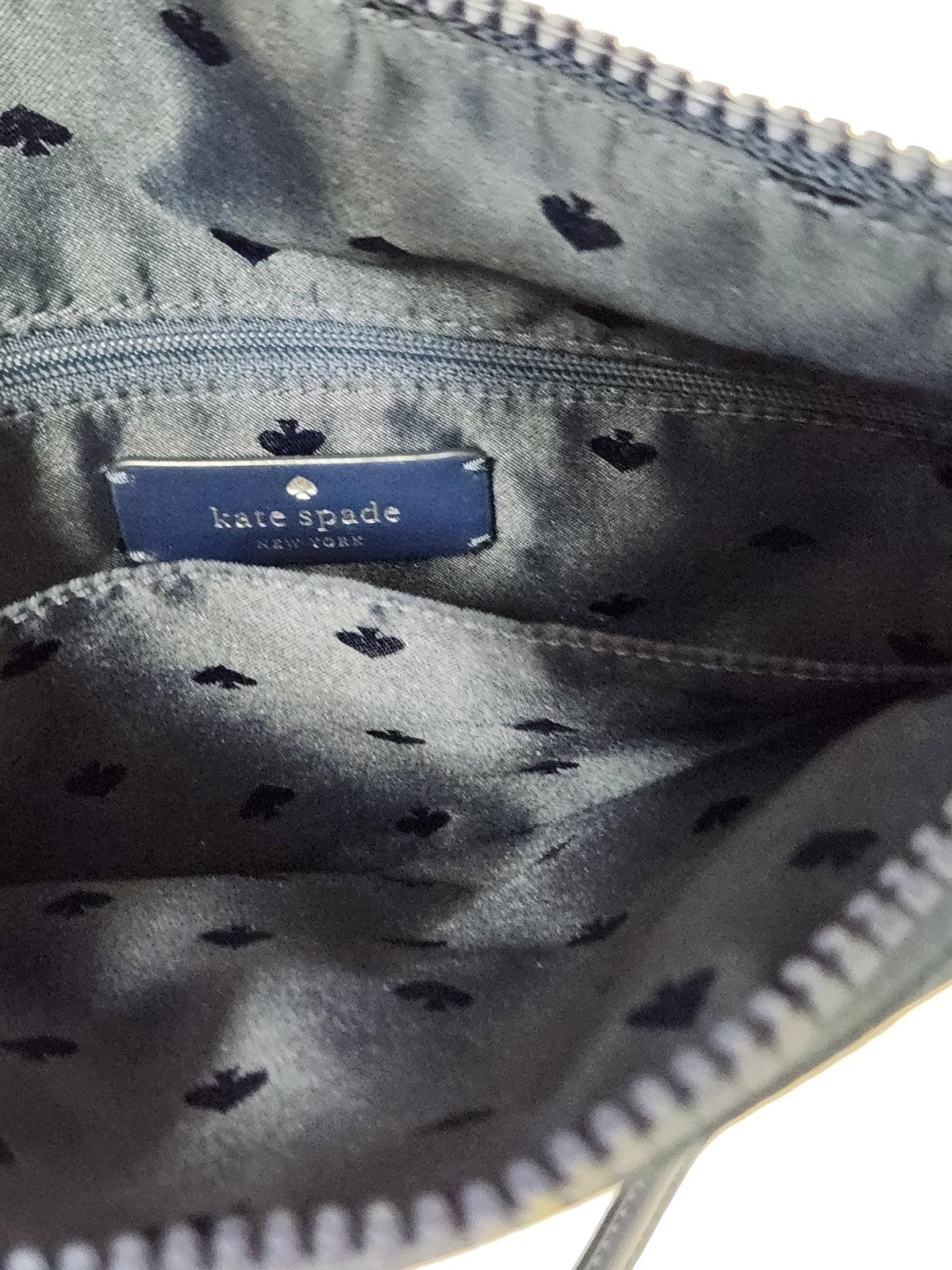 Close-up of the interior of a Kate Spade bag, showing a navy blue brand label sewn onto a gray satin-like lining patterned with small black spade motifs.