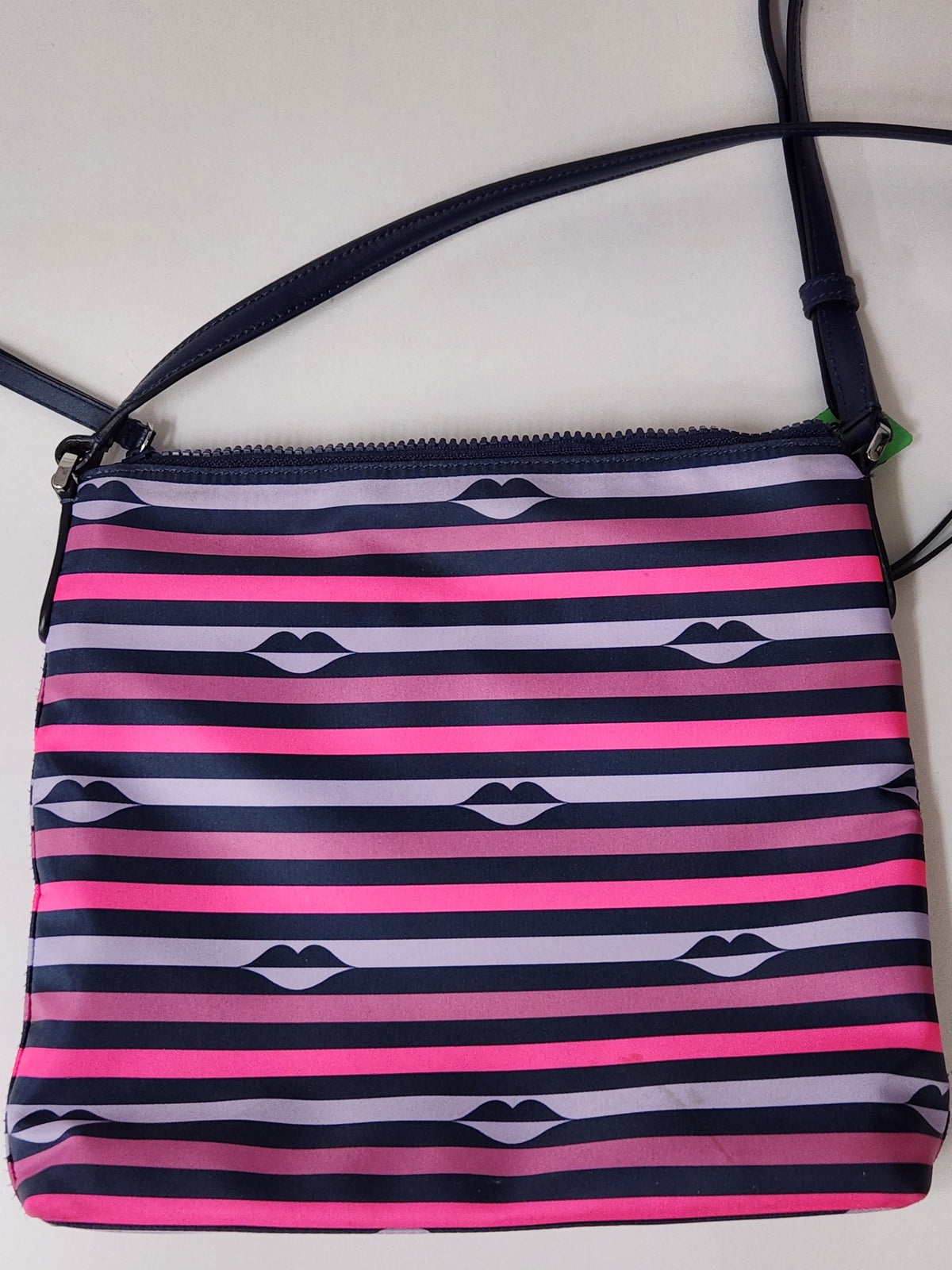 A rectangular crossbody bag with horizontal stripes in navy blue, bright pink, and light lavender, featuring a repeating lip print motif. The bag has a dark navy strap and is photographed against a plain off-white background.