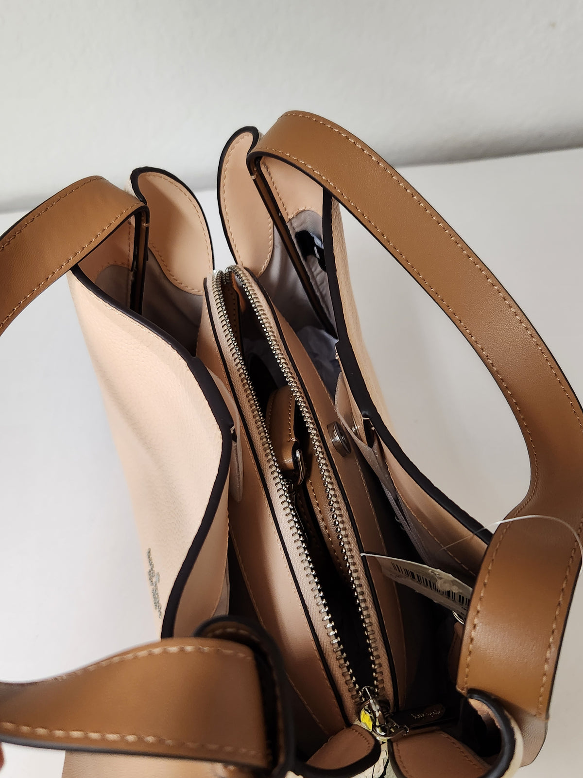 Kate Spade pebbled leather shoulder bag in beige and brown, featuring a top handle, a small top zip pocket, and the brand logo on the front, shown with a retail tag attached against a plain white background.