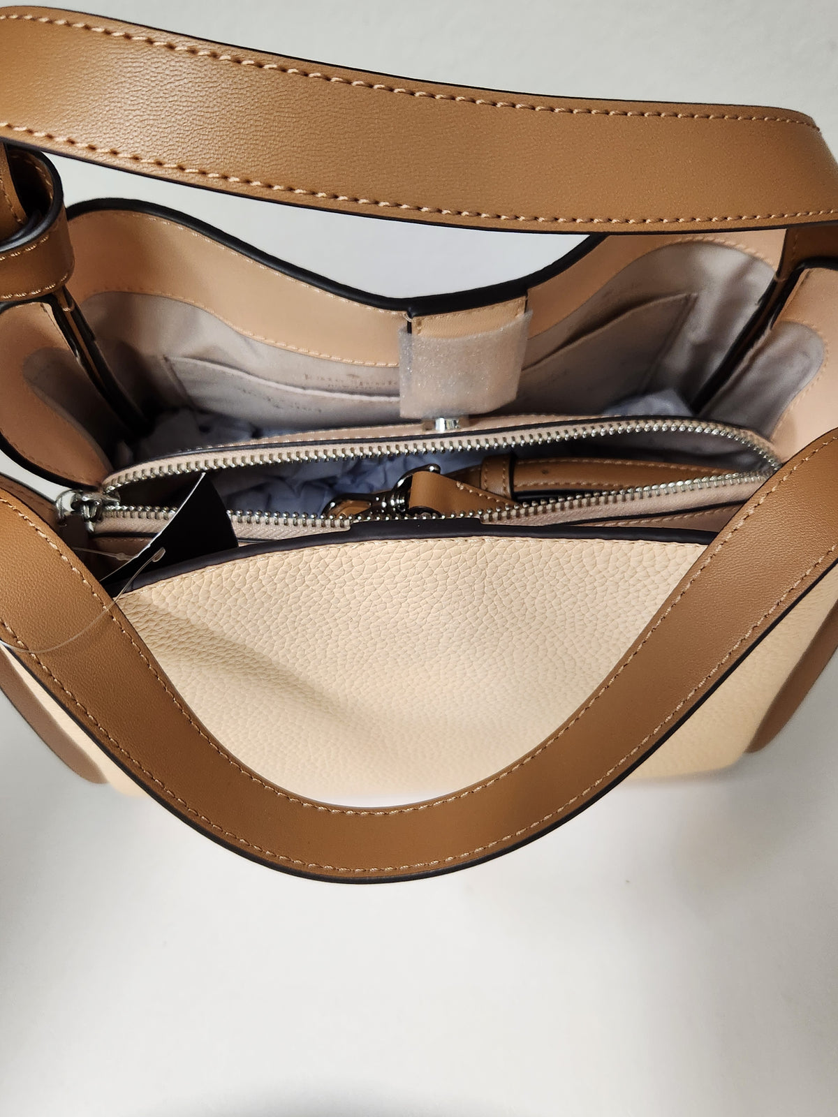 Kate Spade pebbled leather shoulder bag in beige and brown, featuring a top handle, a small top zip pocket, and the brand logo on the front, shown with a retail tag attached against a plain white background.
