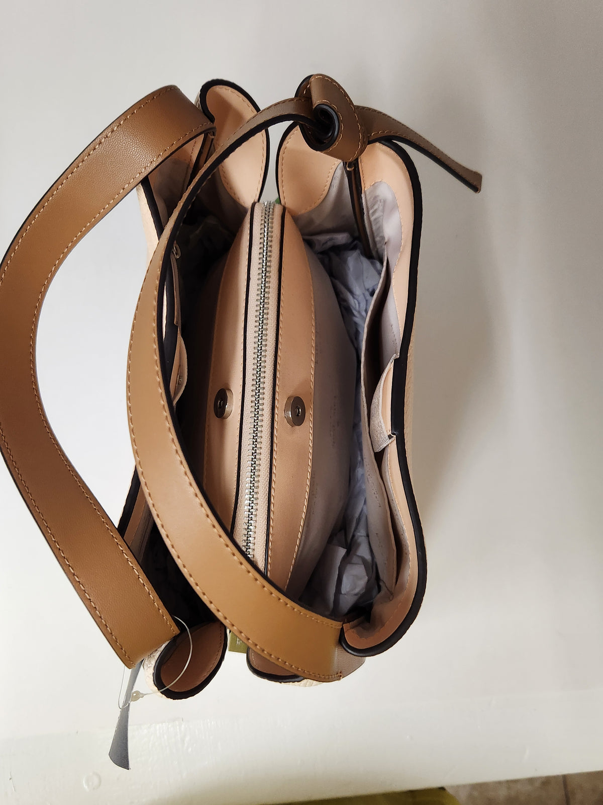 Kate Spade pebbled leather shoulder bag in beige and brown, featuring a top handle, a small top zip pocket, and the brand logo on the front, shown with a retail tag attached against a plain white background.