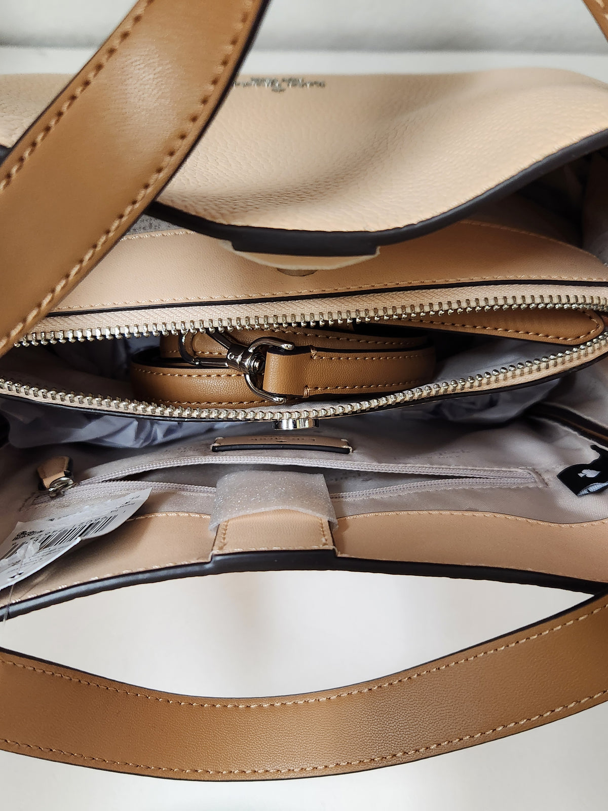 Kate Spade pebbled leather shoulder bag in beige and brown, featuring a top handle, a small top zip pocket, and the brand logo on the front, shown with a retail tag attached against a plain white background.