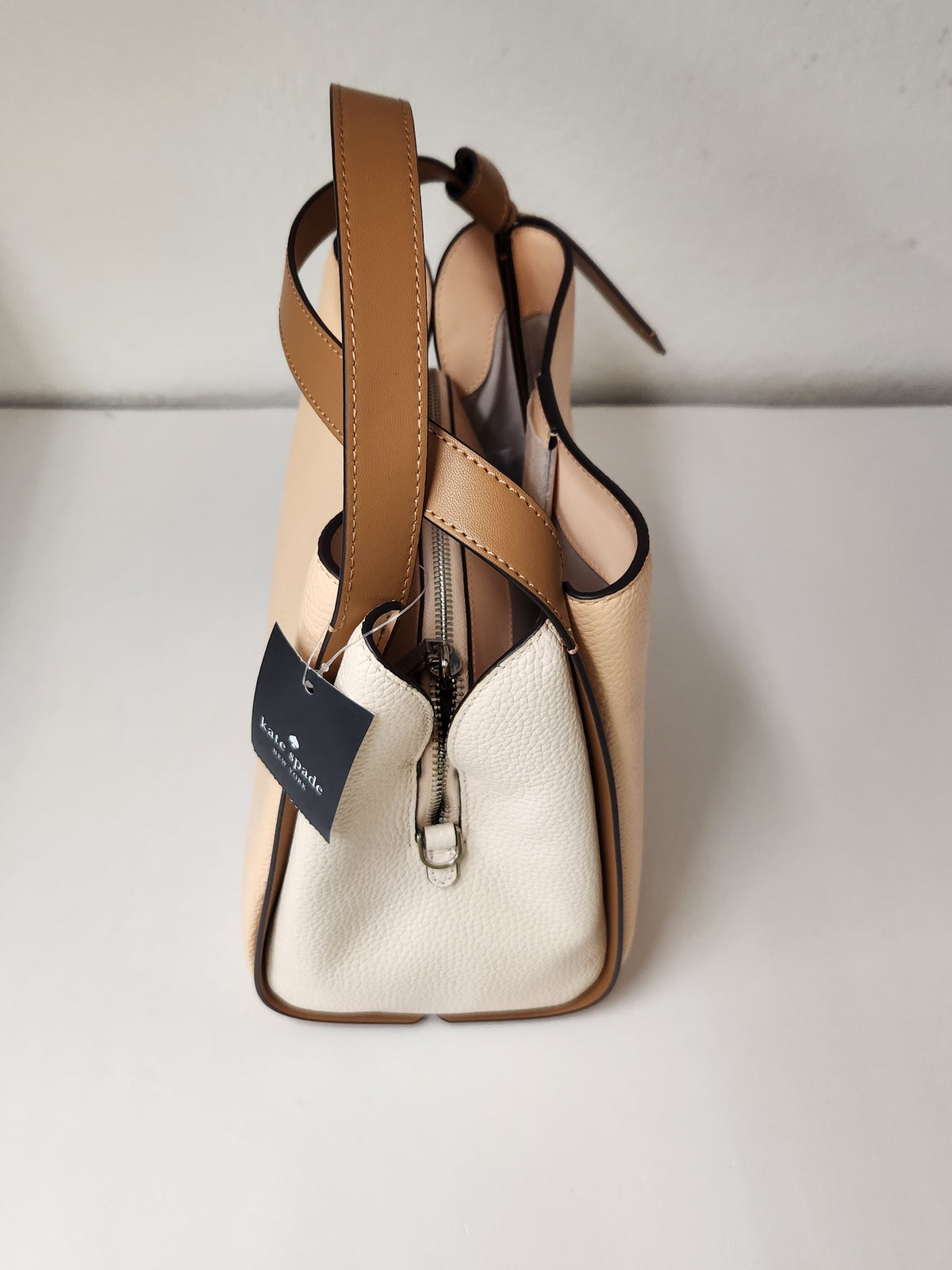 A Kith handbag in beige and off-white pebbled leather with tan leather straps, a silver zipper, and a black Kith hangtag, photographed on a white surface against a white wall.