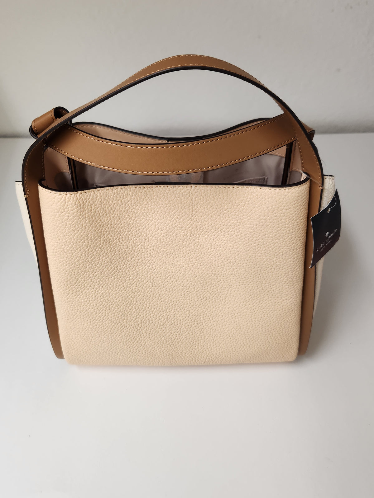A structured pebbled leather handbag in beige and tan, featuring a top handle, a detachable shoulder strap, and a black hangtag on the side, photographed against a neutral light gray background.

