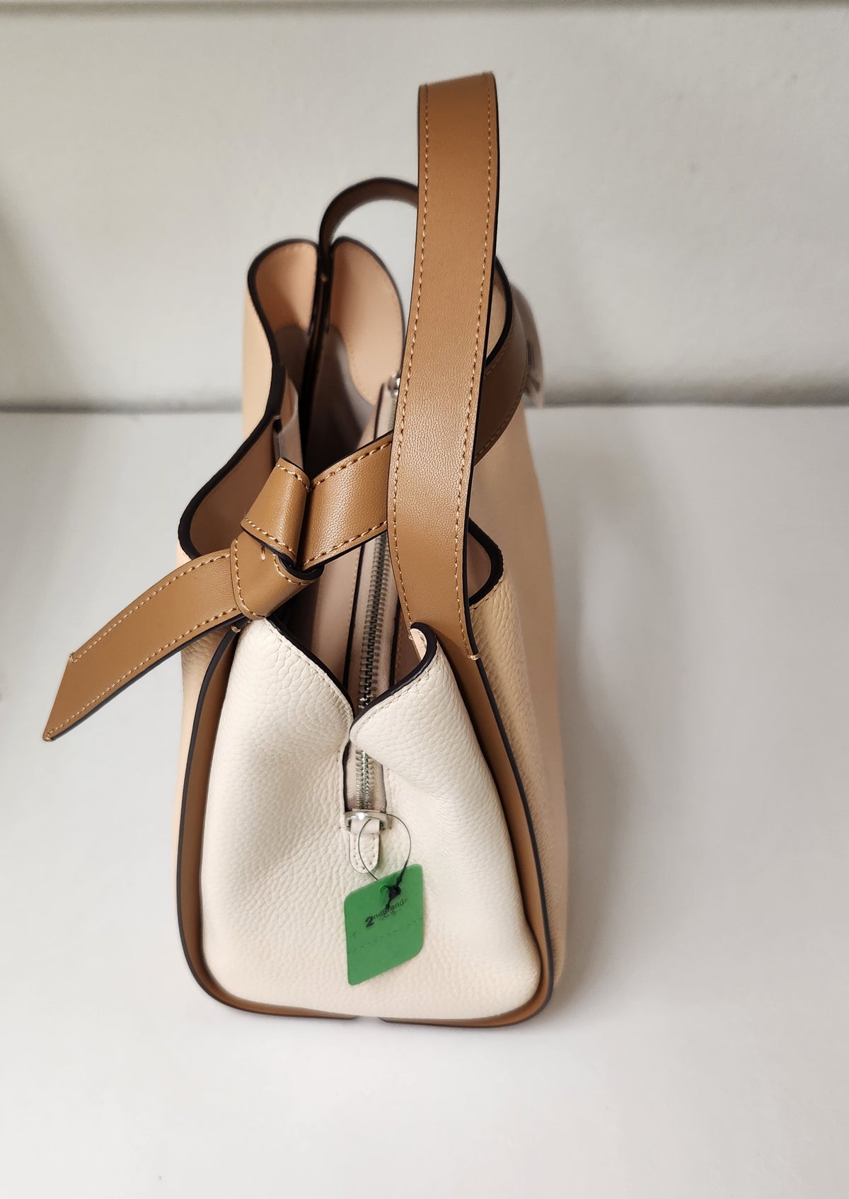A structured pebbled leather handbag in beige and tan, featuring a top handle, a detachable shoulder strap, and a black hangtag on the side, photographed against a neutral light gray background.

