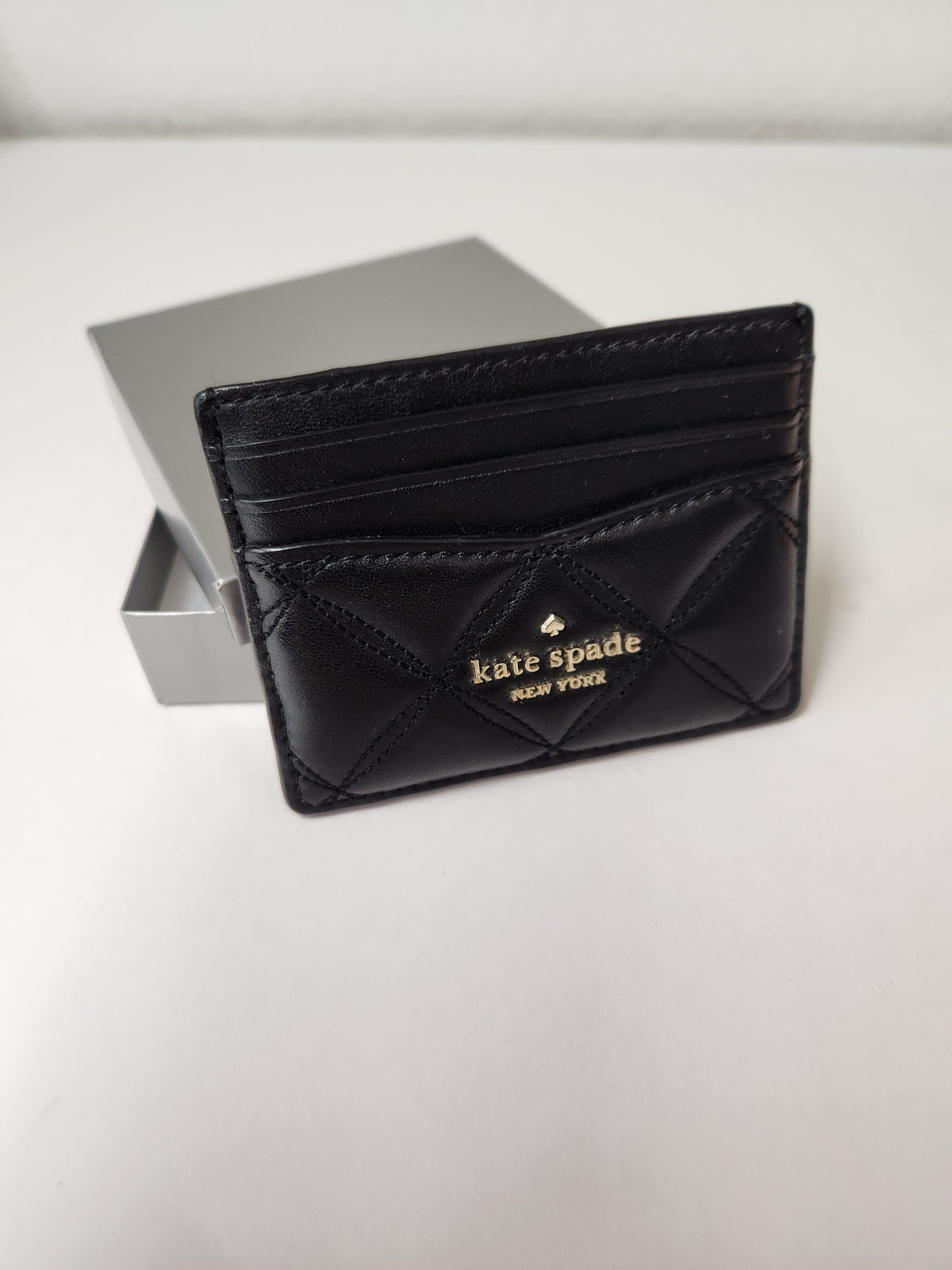 A black leather card holder wallet by Kate Spade New York, featuring a quilted chevron pattern and gold logo, shown next to its silver presentation box on a white surface.