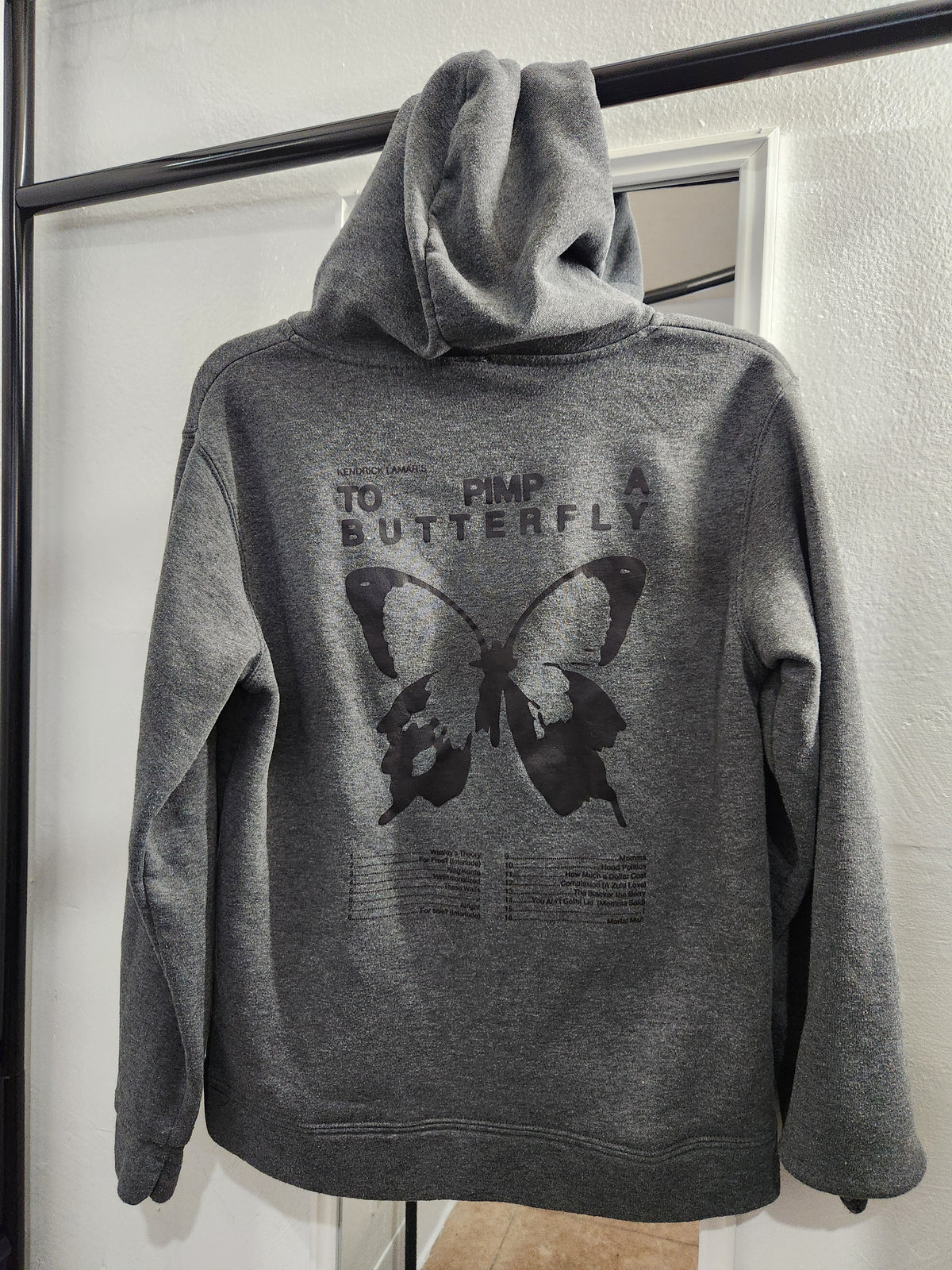 A dark gray heather hoodie hanging on a black clothing rack, showing the back graphic with a butterfly and the text 'TO PIMP A BUTTERFLY'.