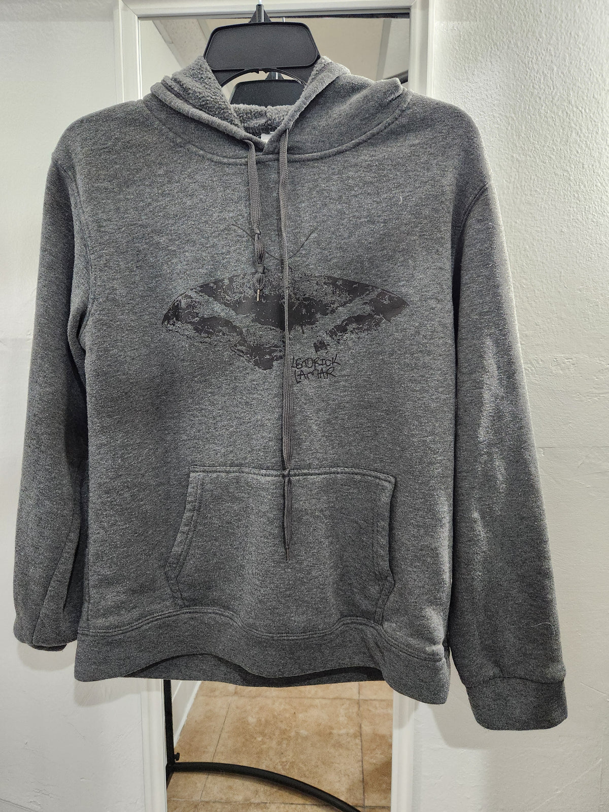A heather gray pullover hoodie with a distressed black graphic on the chest, hanging on a black hanger against a white door frame.