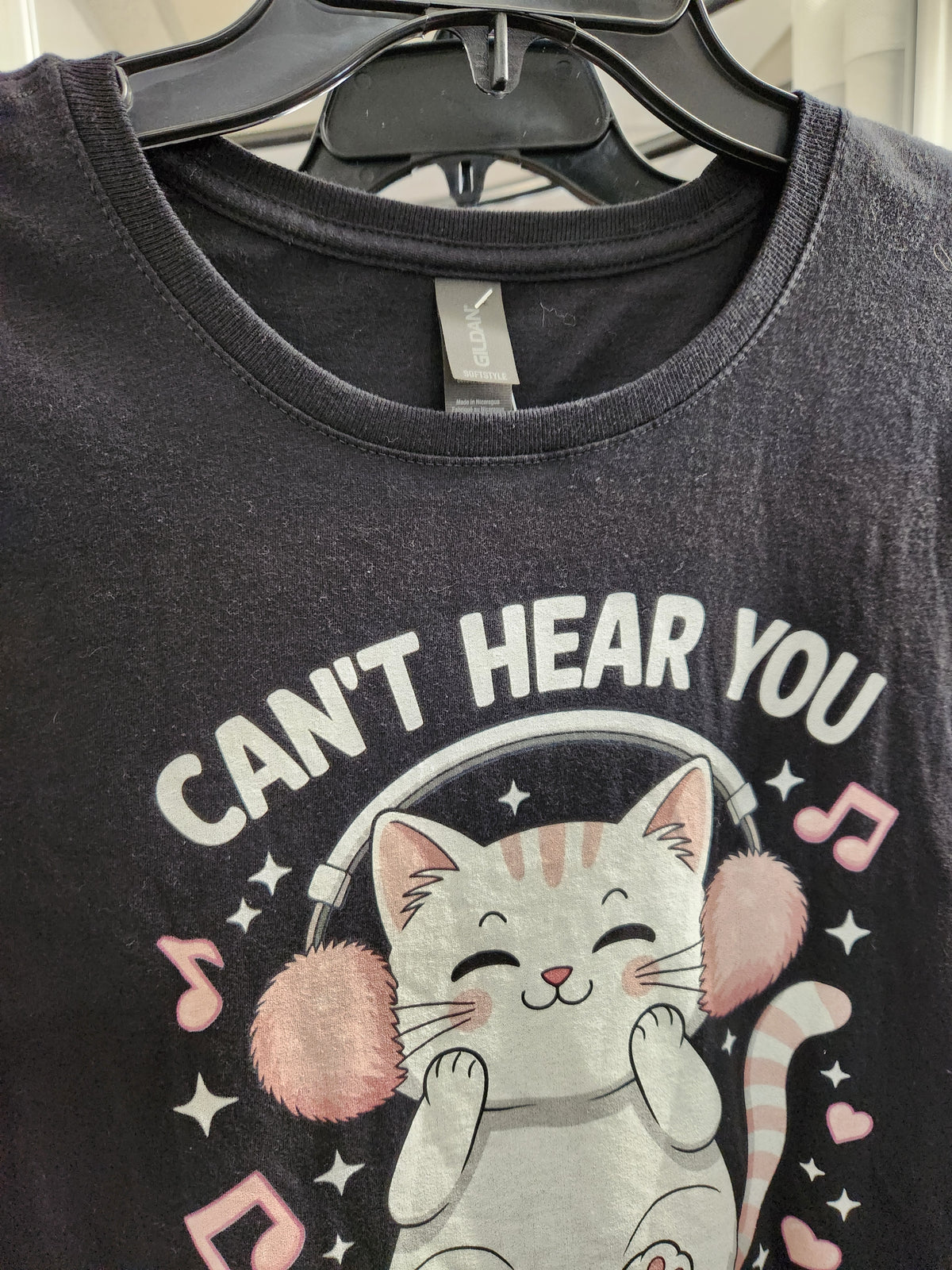 A dark charcoal gray graphic t-shirt hanging on a black hanger. The shirt features a cartoon illustration of a white cat wearing pink earmuffs with the text 'CAN'T HEAR YOU' in white capital letters.