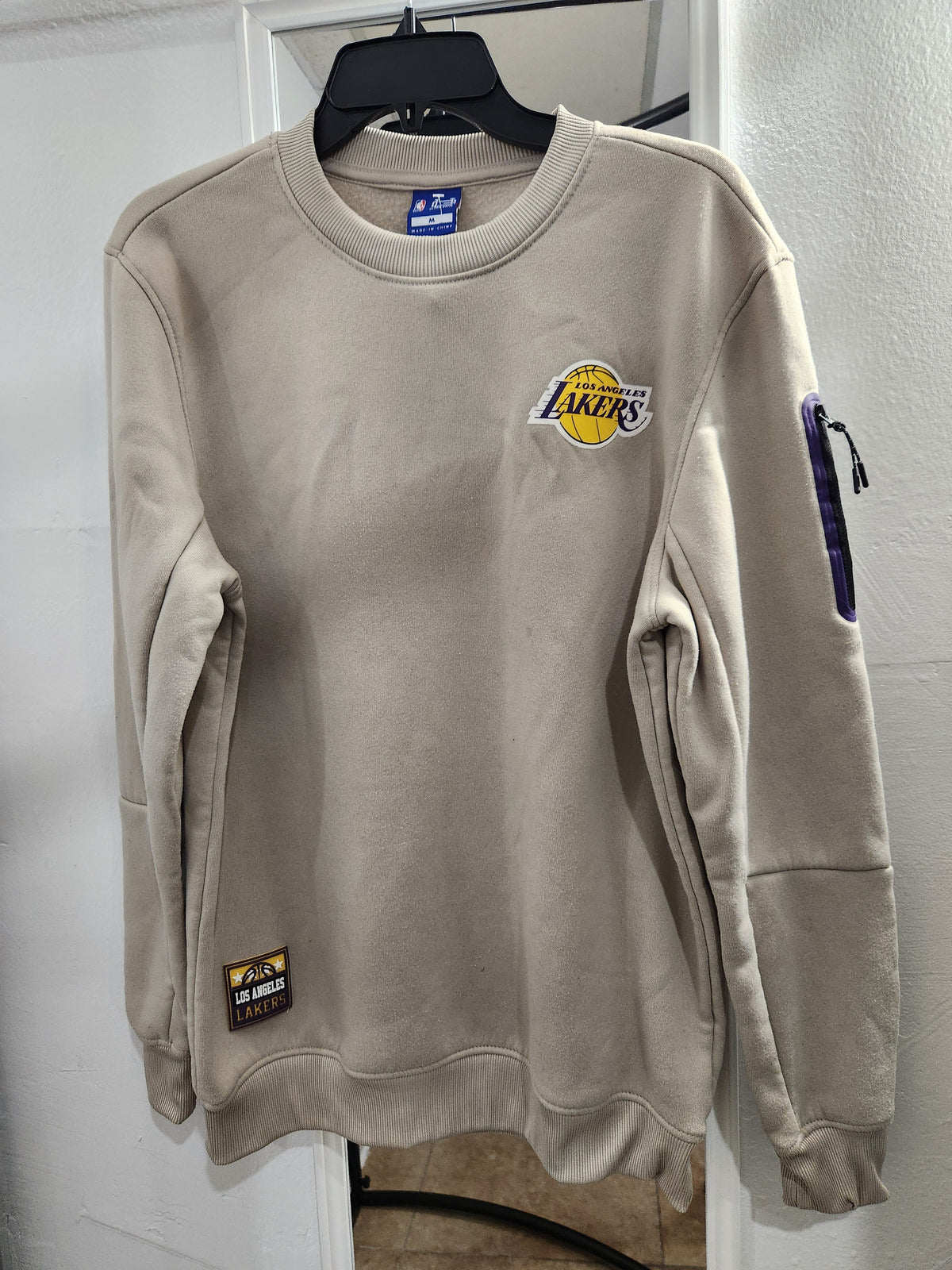 A beige Los Angeles Lakers crewneck sweatshirt hanging on a black hanger against a white door frame. The sweatshirt features the Lakers logo on the left chest, a small brand tag near the bottom left hem, and a purple graphic detail on the right sleeve.