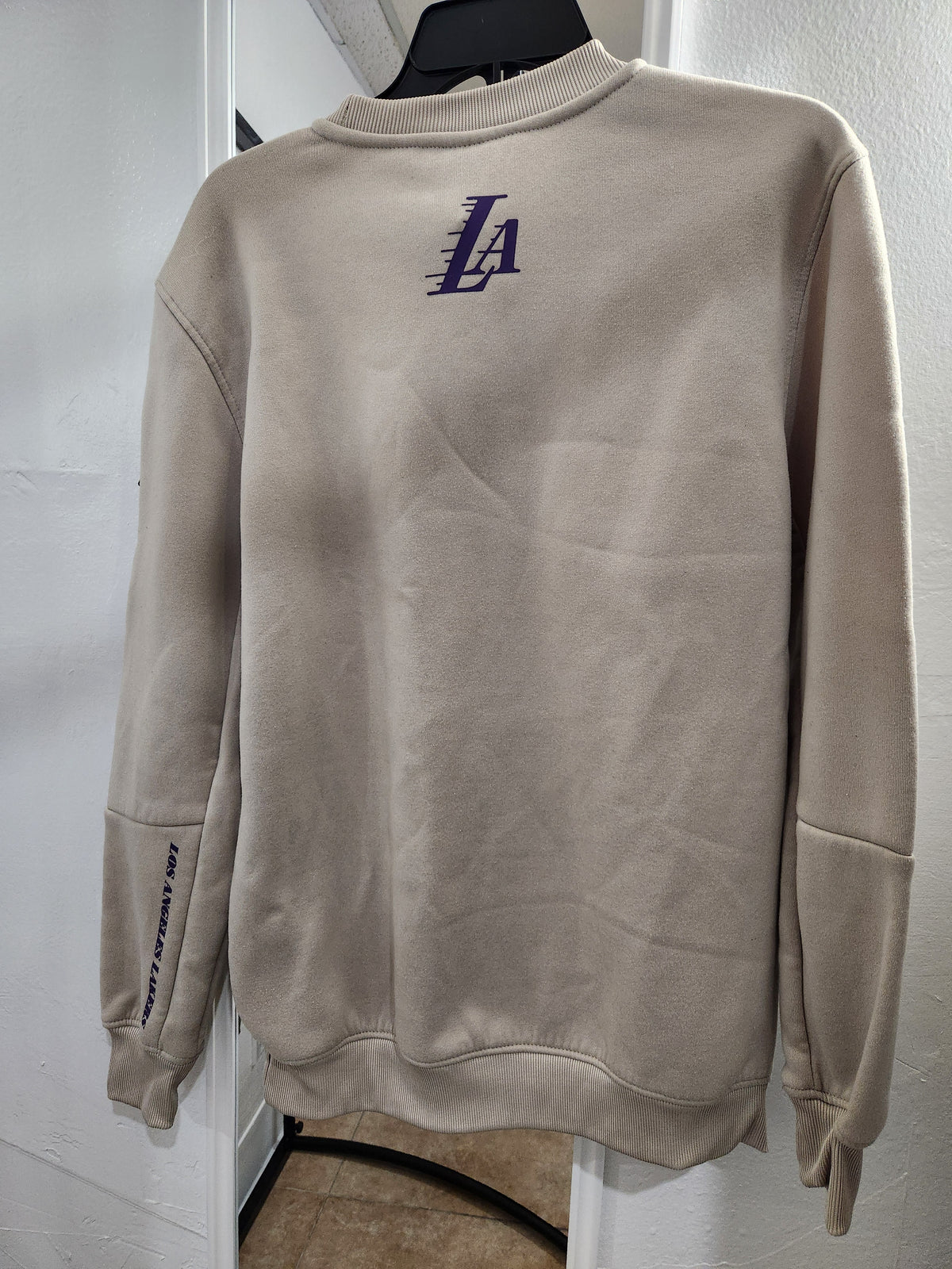 A beige crewneck sweatshirt displayed on a black hanger against a white wall. The sweatshirt features a purple stylized 'LA' logo on the chest and 'LA SPORTS' printed vertically on the left sleeve.