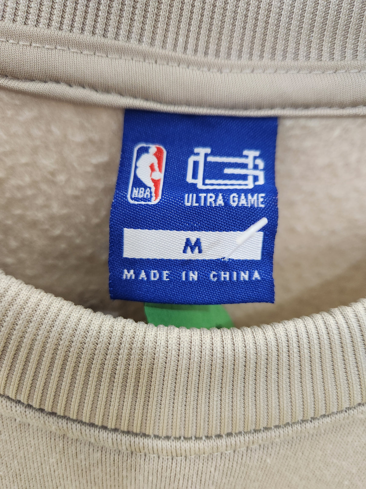 Close-up of the inside neck label of a beige ribbed sweatshirt. The blue NBA Ultra Game tag shows size M and 'Made in China' with the NBA logo visible.