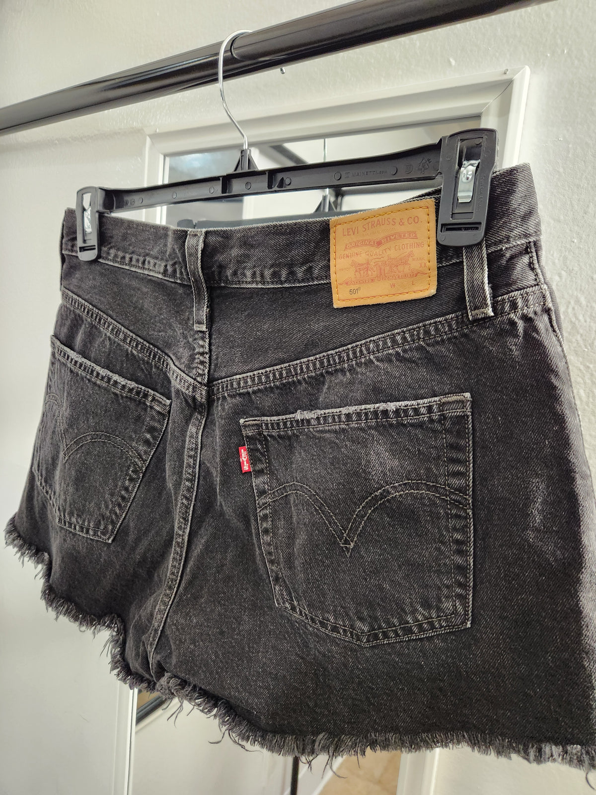 Back view of a pair of dark gray washed denim shorts hanging on a black hanger with clips, showing the leather patch and red tab on a white door frame.