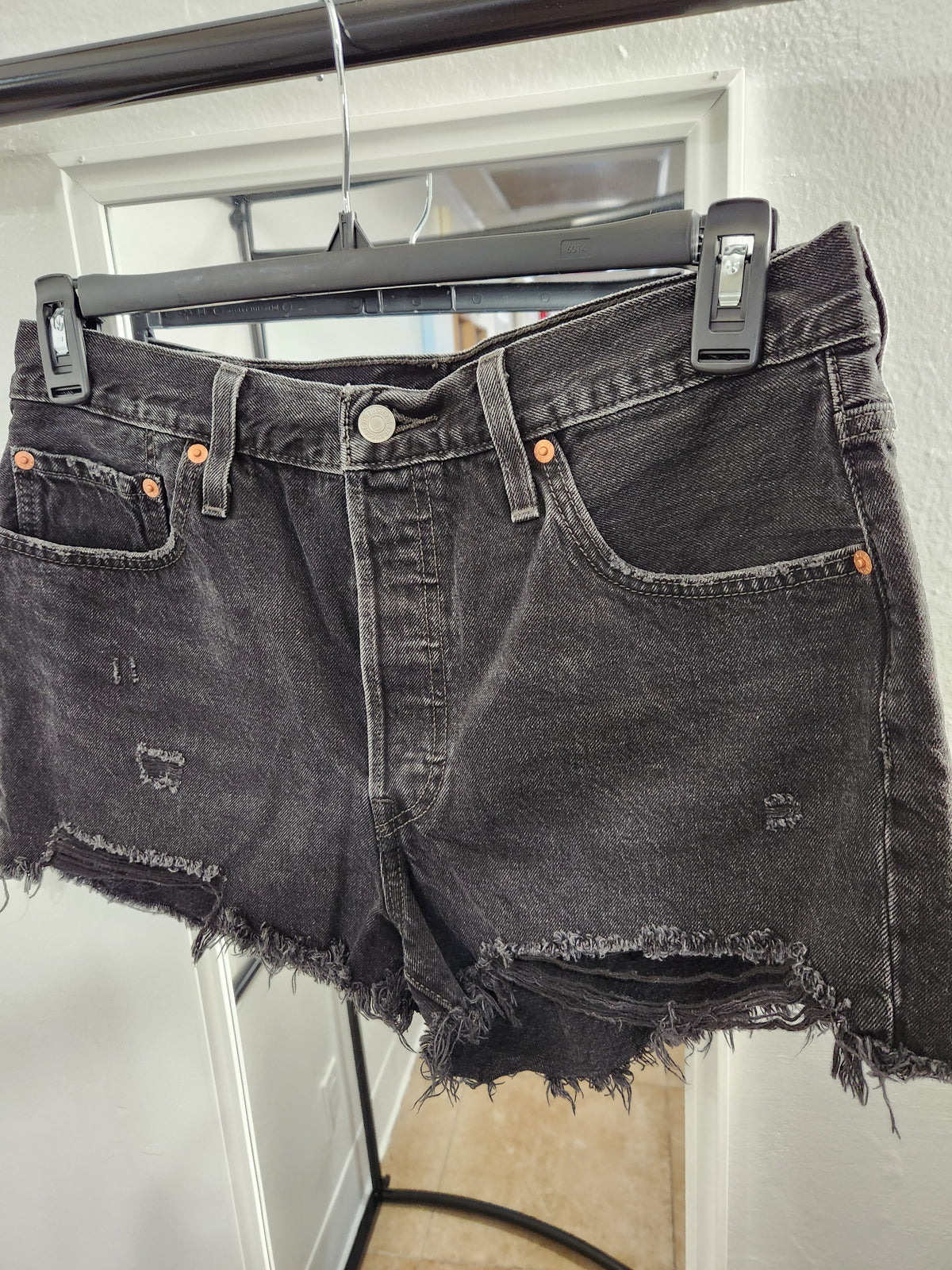 Black washed denim shorts with a distressed, frayed hem and orange-brown copper-toned rivets, hanging on a black hanger.