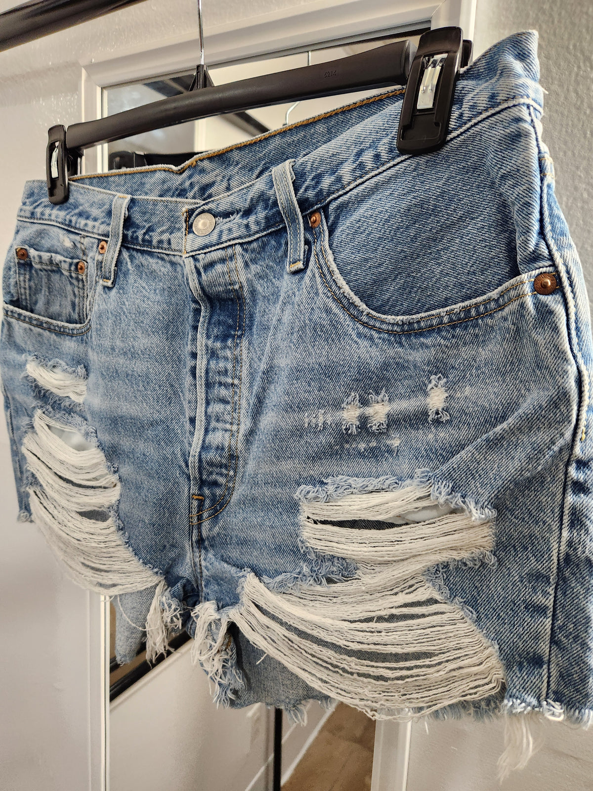 Light wash blue denim shorts with heavy distressing and ripped details, hanging on a black hanger.