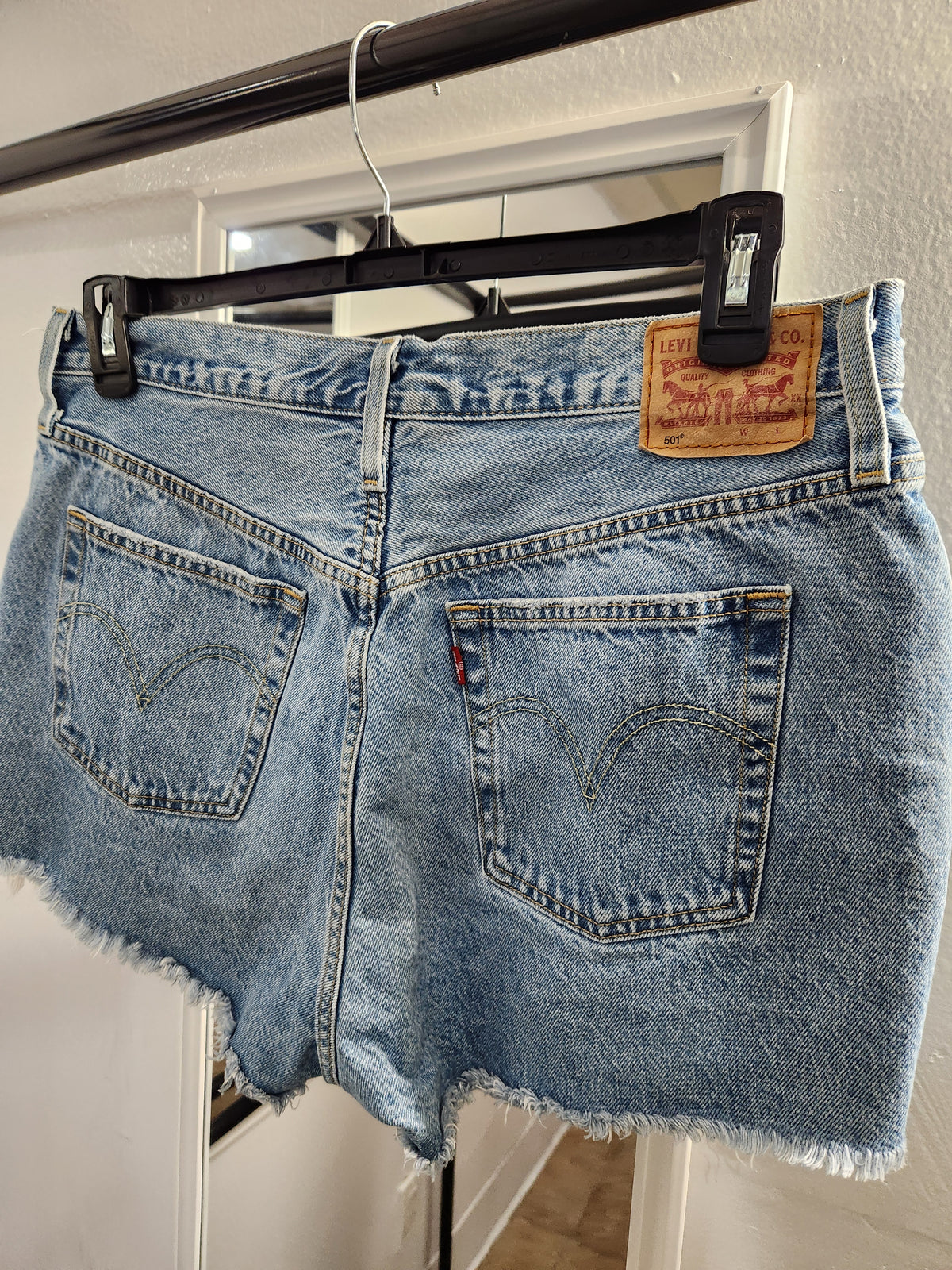 Light blue Levi's denim shorts with a raw, frayed hem, hanging on a black hanger with clips against a white door frame.