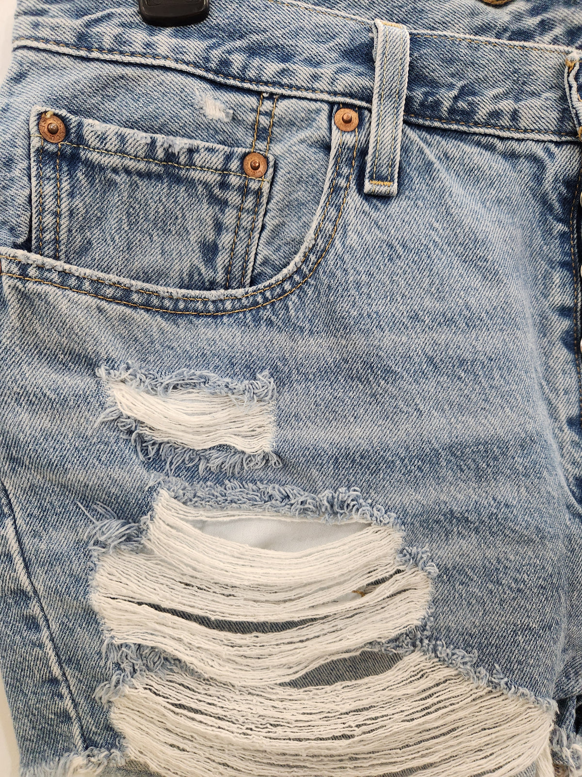 Close-up of light blue distressed denim shorts showing the front pocket, belt loops, rivets, and large ripped areas with frayed white cotton detailing.
