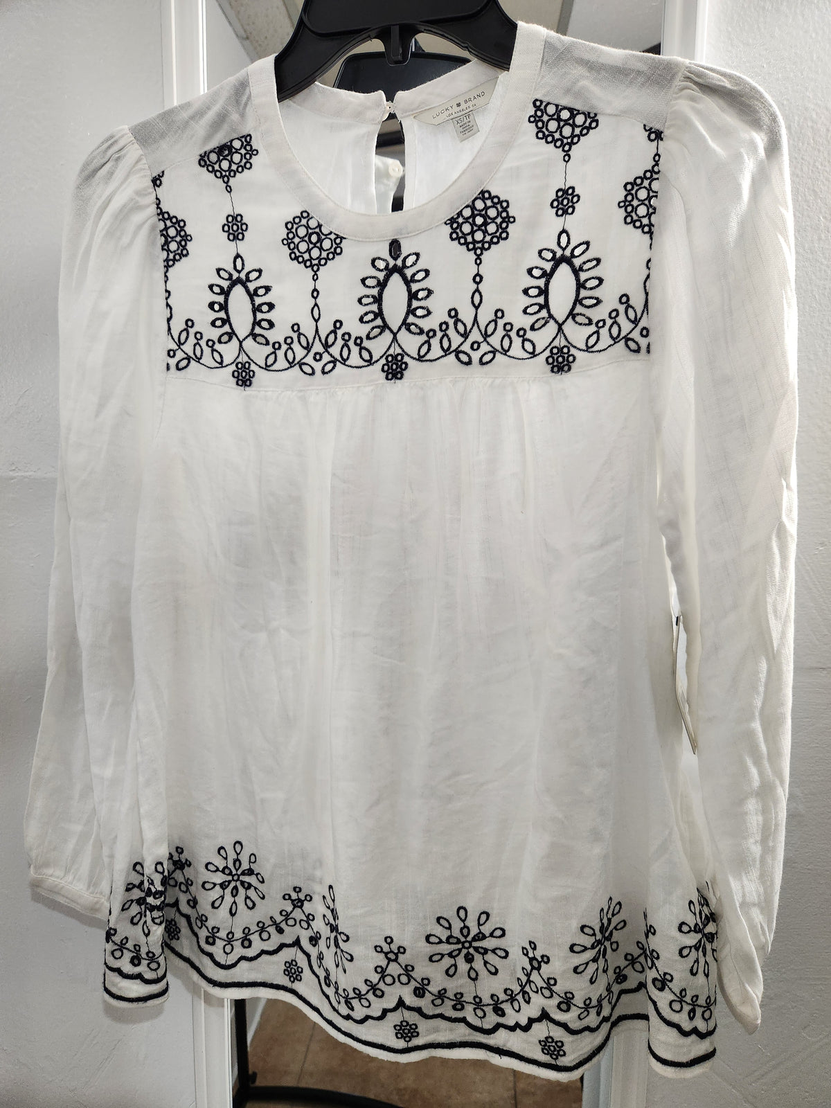 A white long-sleeve blouse with black embroidered floral and geometric patterns across the chest and along the hem, displayed on a black hanger against a white paneled background.
