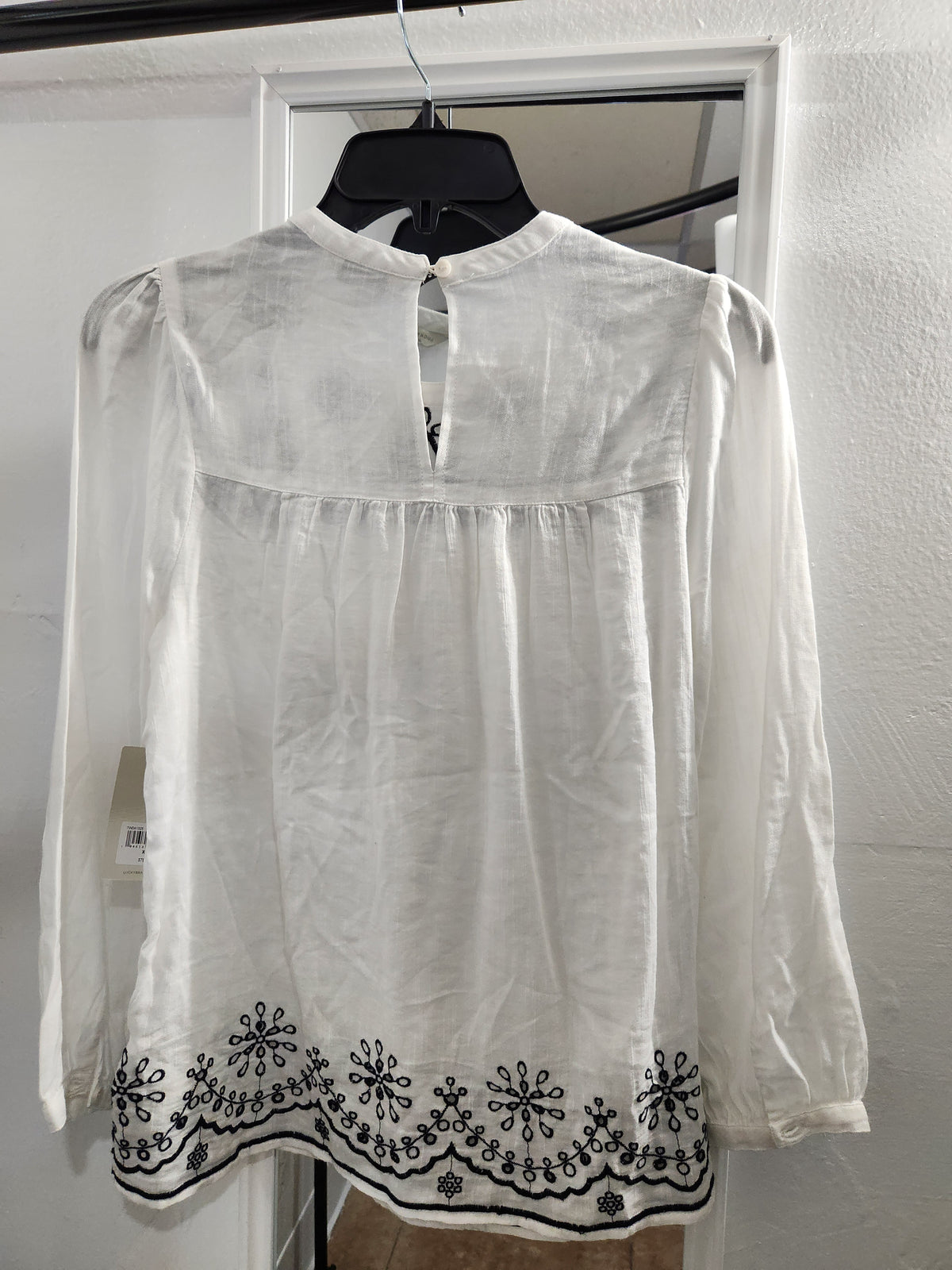 A white long-sleeve blouse with black floral embroidery along the hem, hanging on a black hanger against a white door and textured wall.