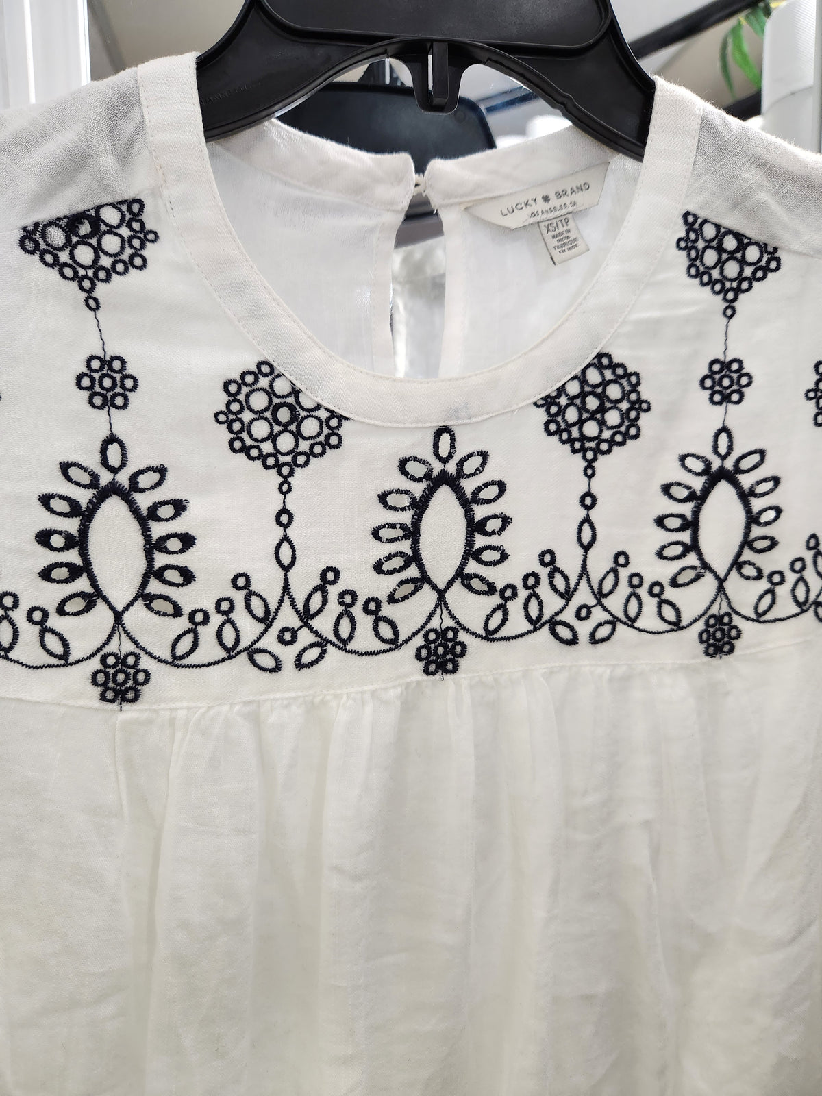 White cotton blouse with black floral embroidery across the chest, hanging on a black hanger. The brand label reads 'LUCKY & BEAR'.