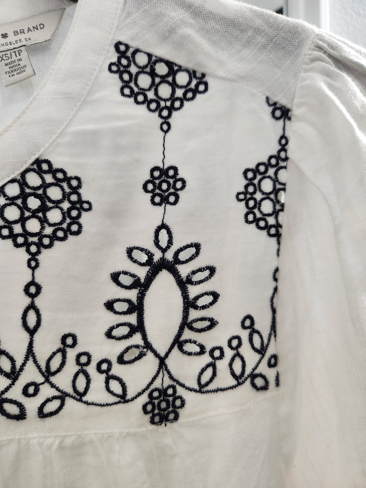 Close-up of a white embroidered blouse with black floral and geometric embroidery on the chest, showing part of the collar and a brand tag.