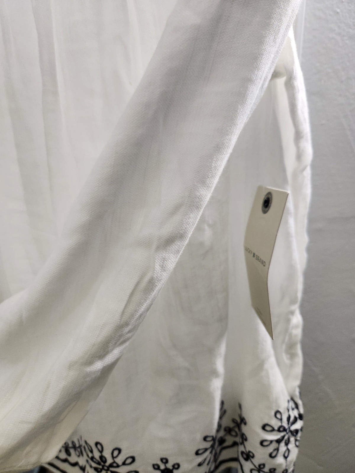 A close-up of a white fabric garment or textile with black embroidered floral border detailing near the bottom and a small paper hangtag attached.
