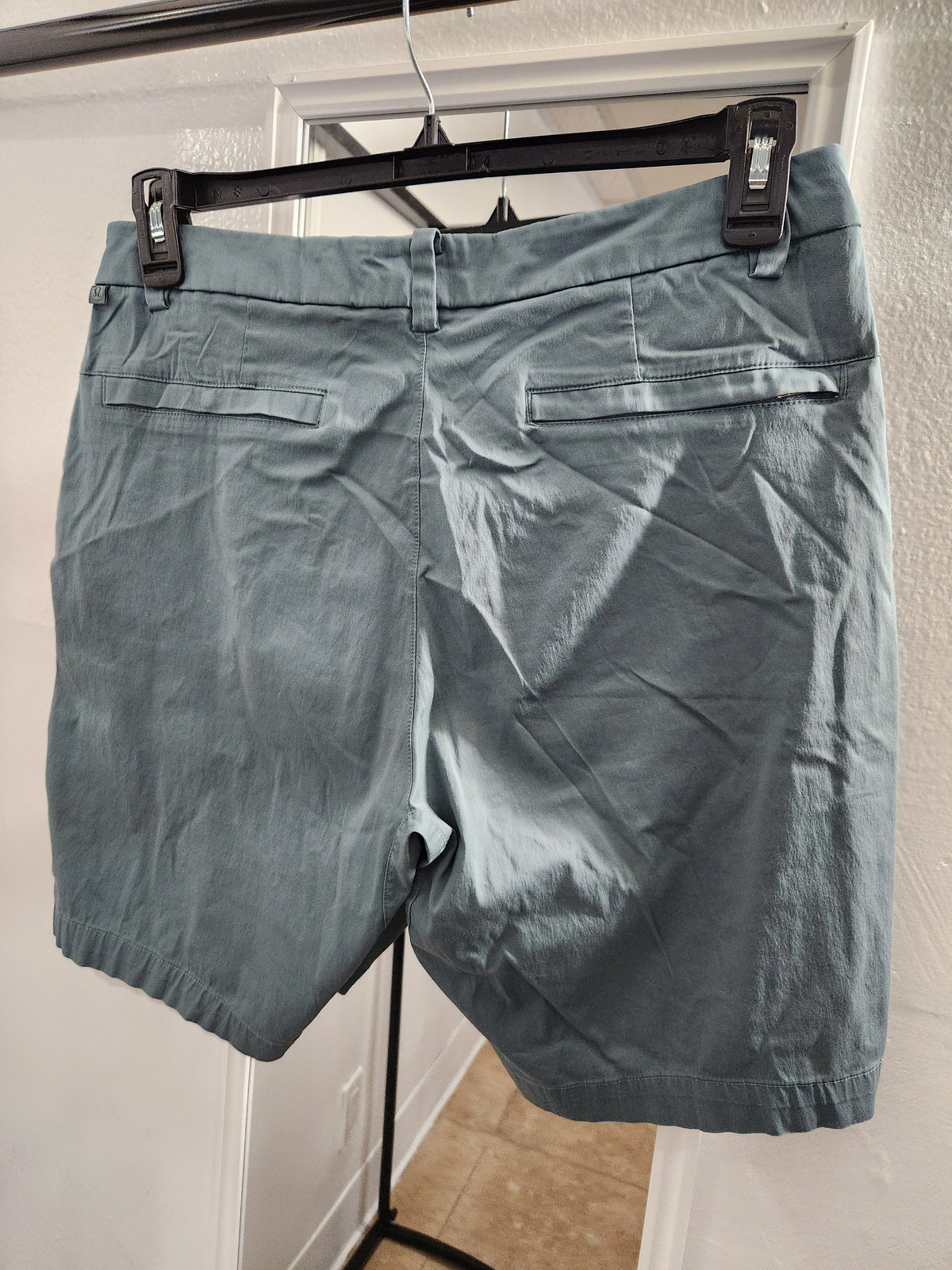 A pair of slate blue-gray shorts hanging from a black hanger on a door frame. The back of the shorts features two welt pockets and a button closure.