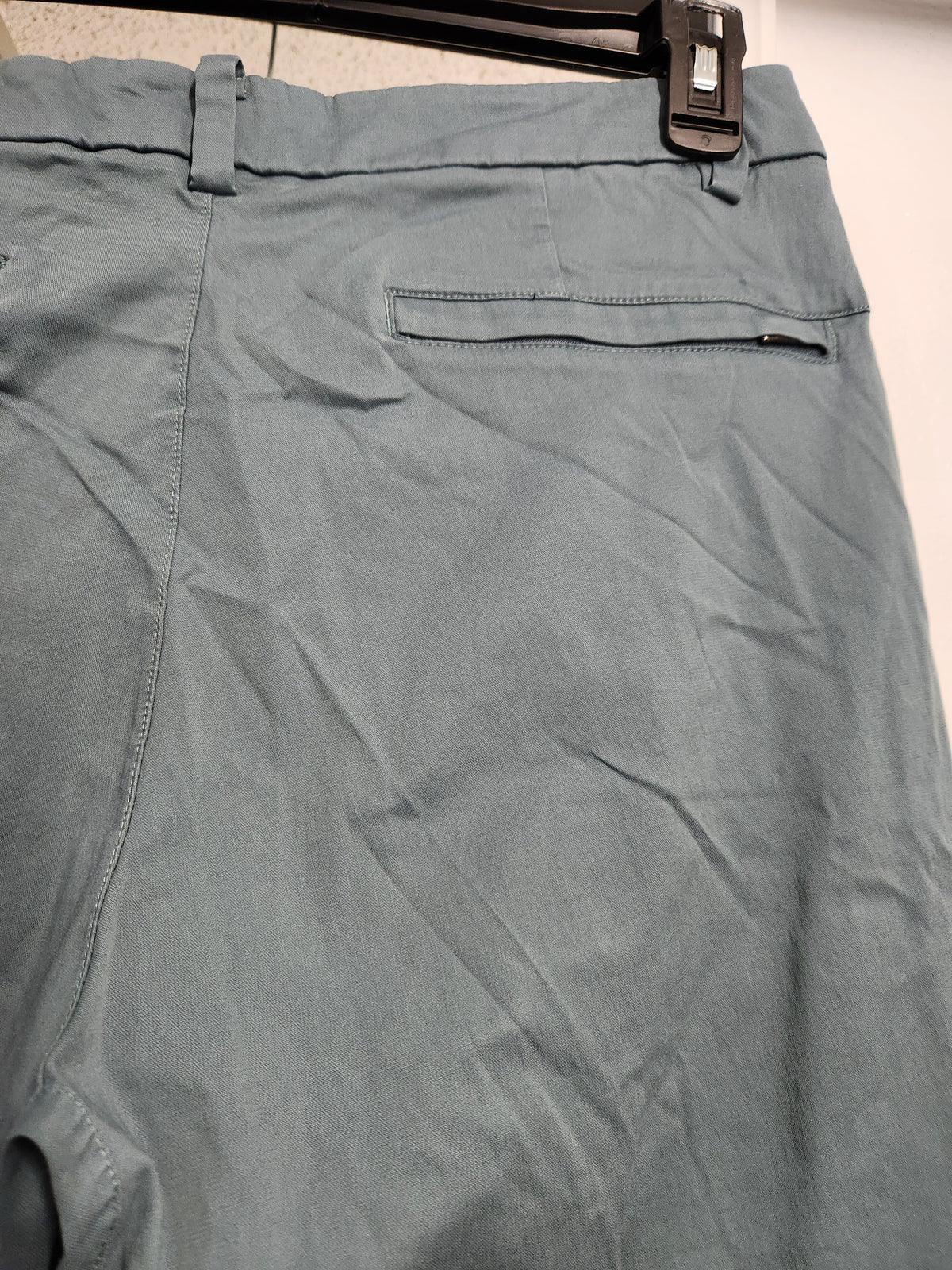 Close-up of the back waistband and upper leg area of a pair of light blue-gray chino pants hanging on a black hanger against a plain light background.