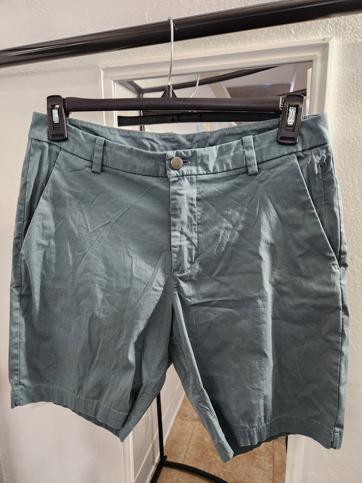 A pair of muted blue-gray chino shorts hanging from a black hanger on a clothing rack, positioned in front of a white door frame.