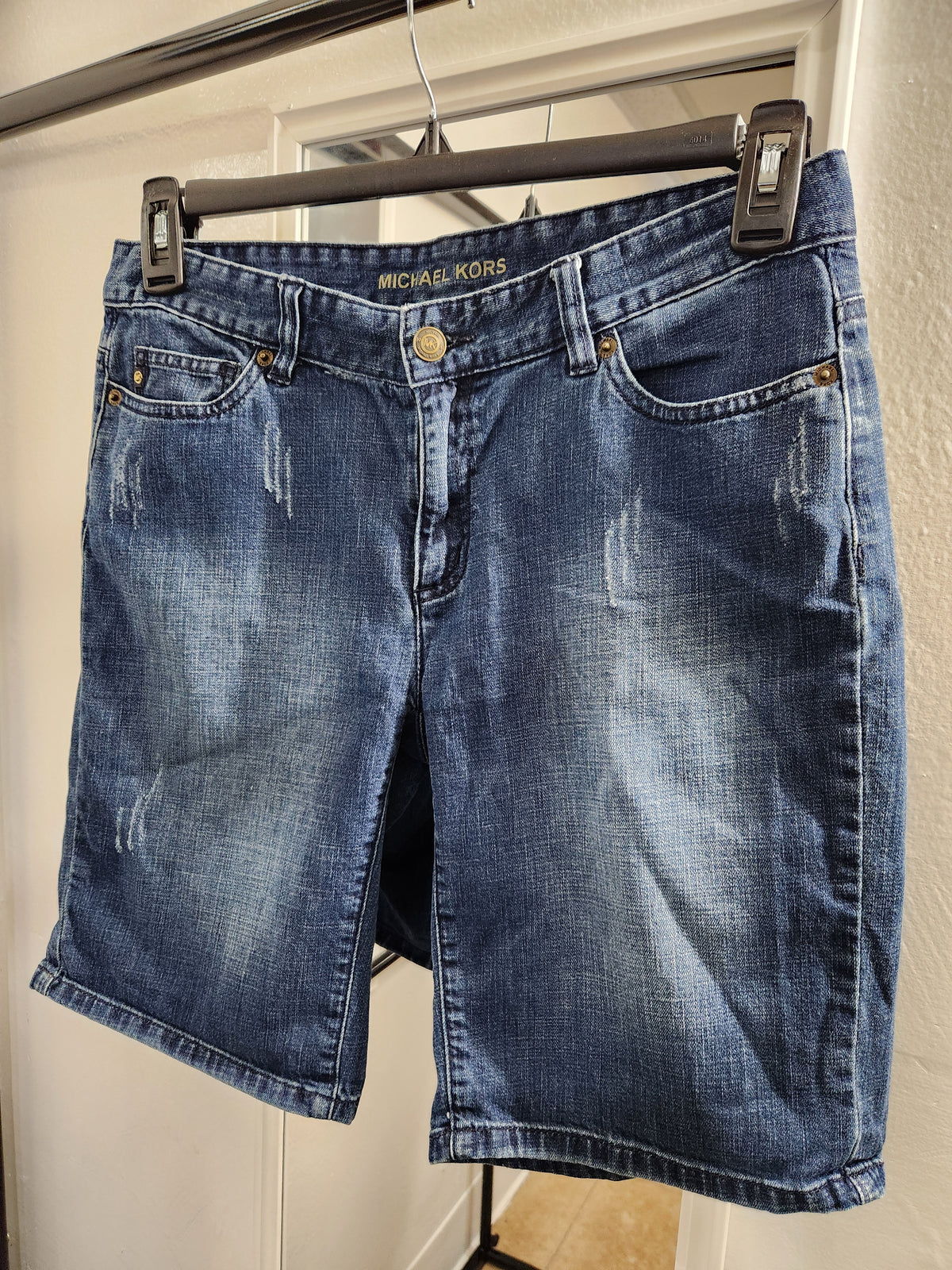 A pair of blue distressed denim shorts with 'MICHAEL KORS' printed in gold on the waistband, hanging from a black hanger against a white door.
