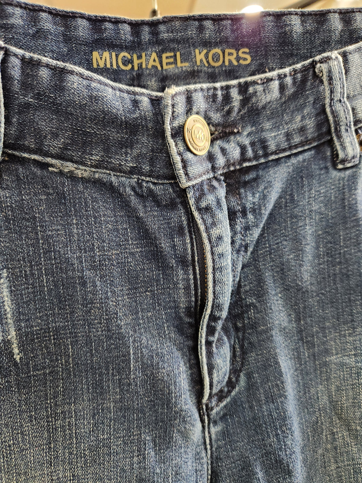 Close-up of blue denim jeans showing the waistband with 'MICHAEL KORS' embroidered in gold, a gold button, and detailed stitching.