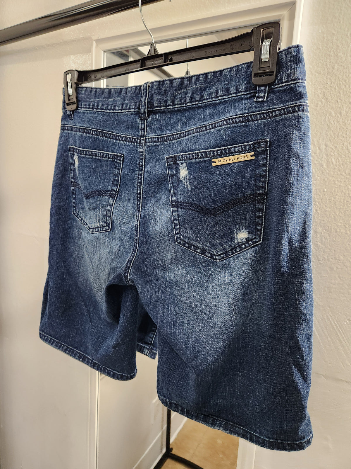 Blue denim shorts with a faded wash, hanging on a black hanger against a white wall. The back of the shorts features two pockets with distressed details and a small gold-tone brand label on the right pocket.