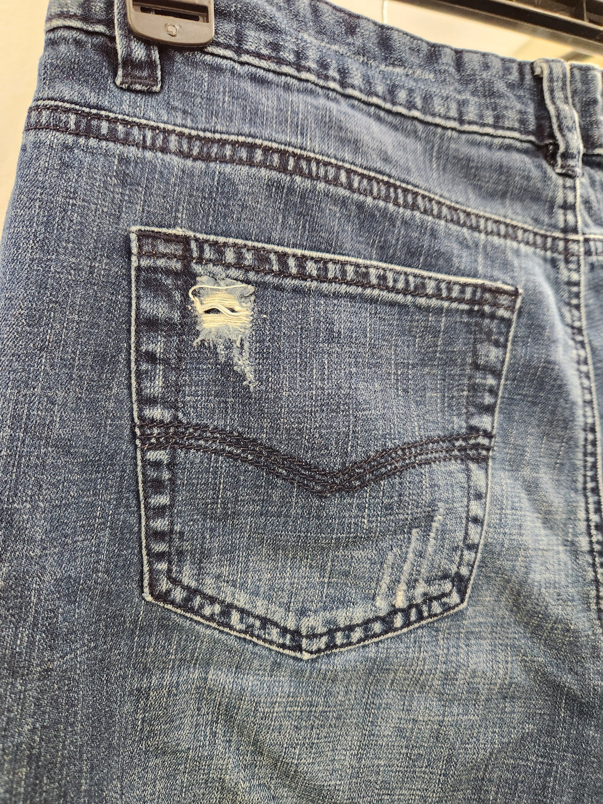 Close-up of the back pocket of light blue denim jeans with a distressed rip on the pocket and dark contrast stitching.