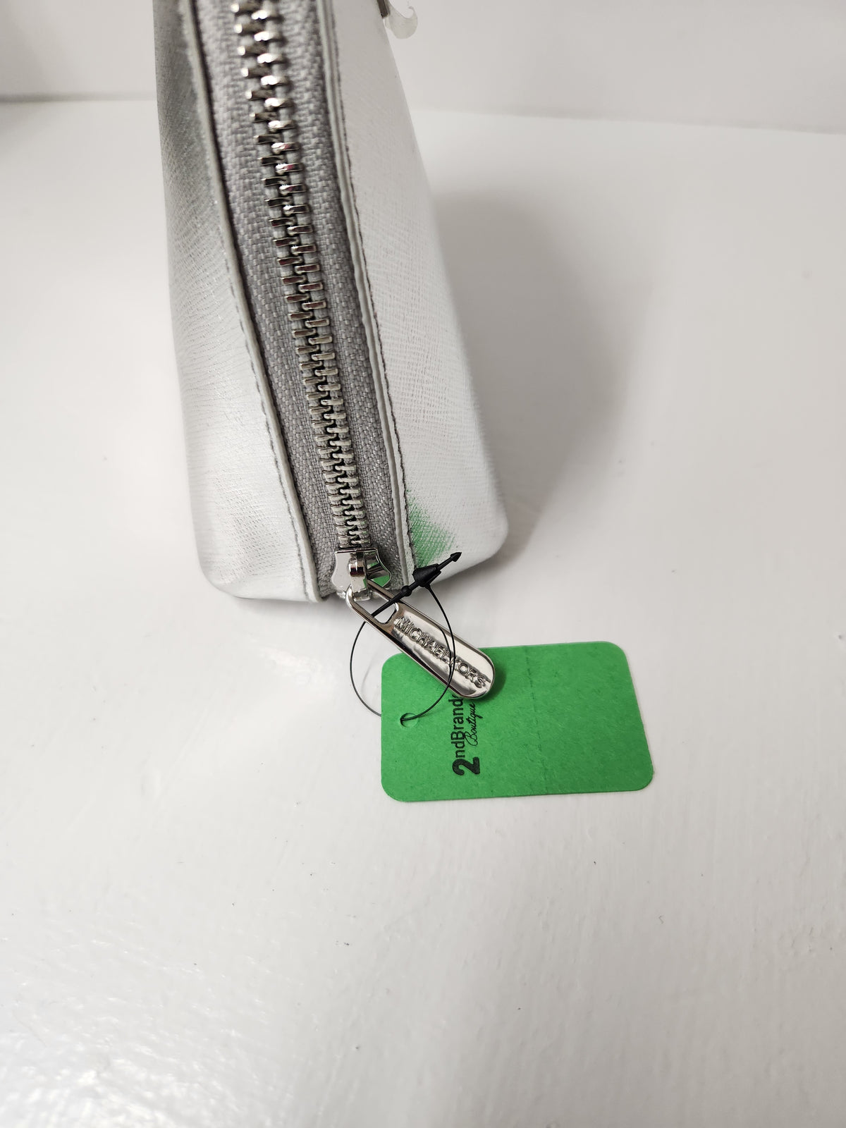 A close-up of a silver metallic zippered pouch with a green paper tag attached, photographed on a white surface.
