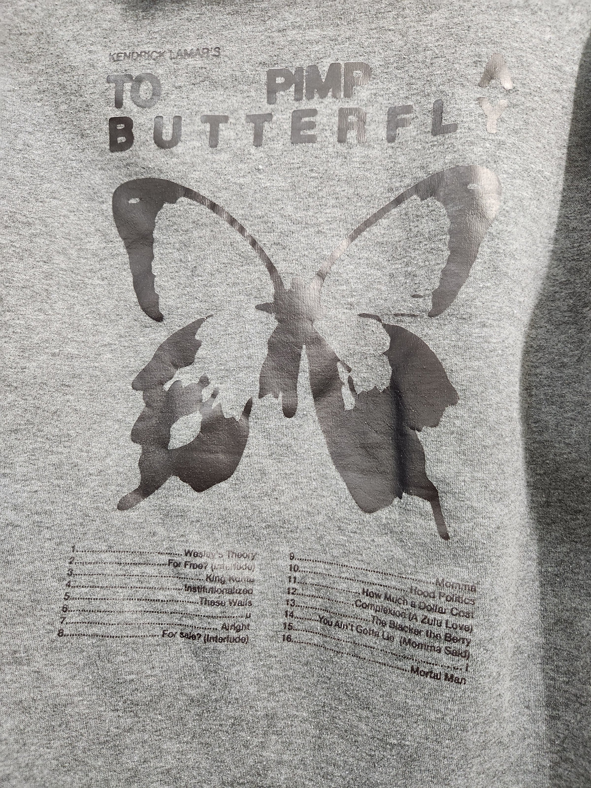 Close-up of a heather gray t-shirt featuring a dark gray graphic print of a butterfly silhouette with the text 'TO PIMP BUTTERFLY' and a tracklist below.
