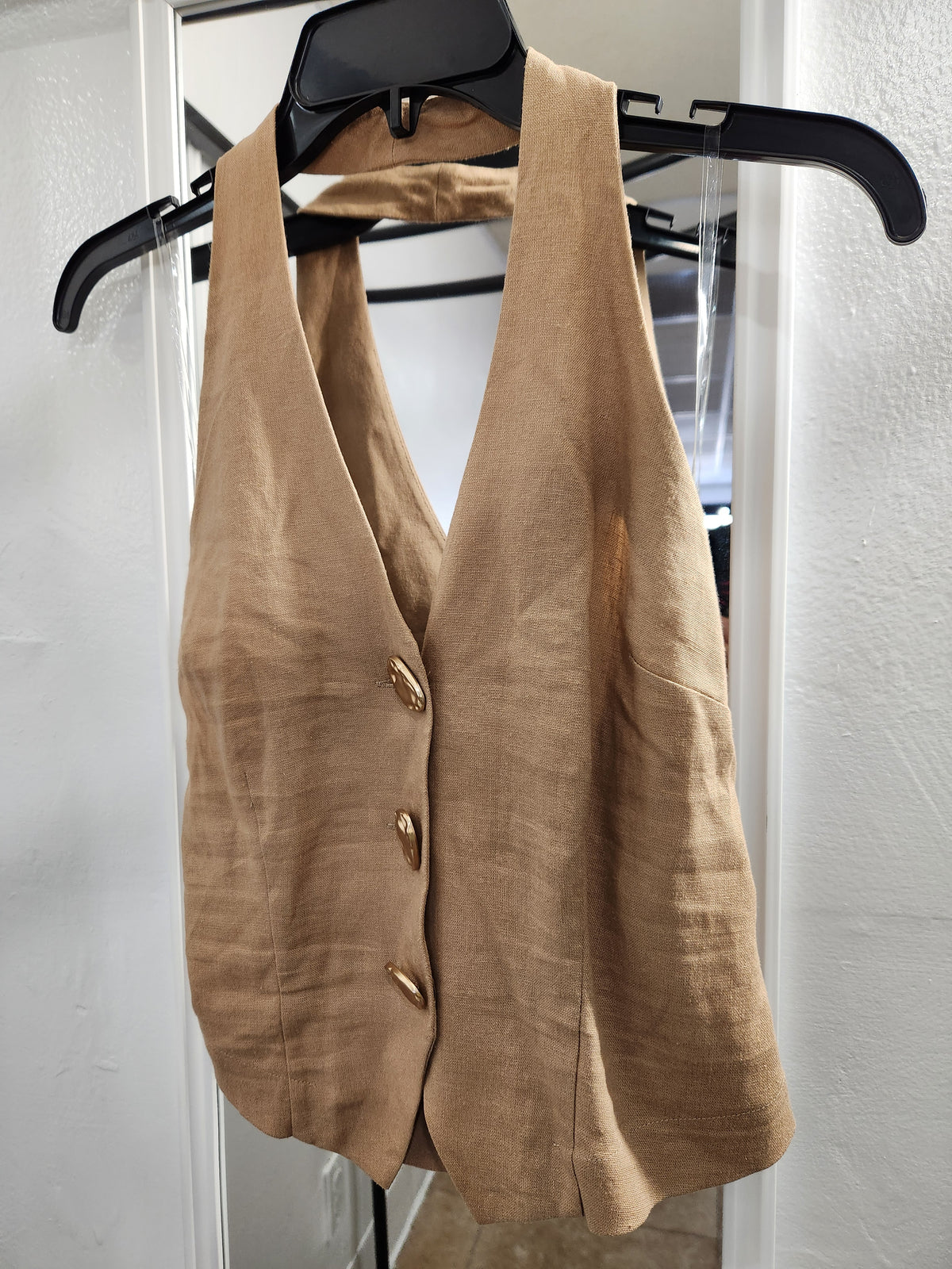A sleeveless tan linen vest with three gold buttons, hanging on a black hanger against a white wall.