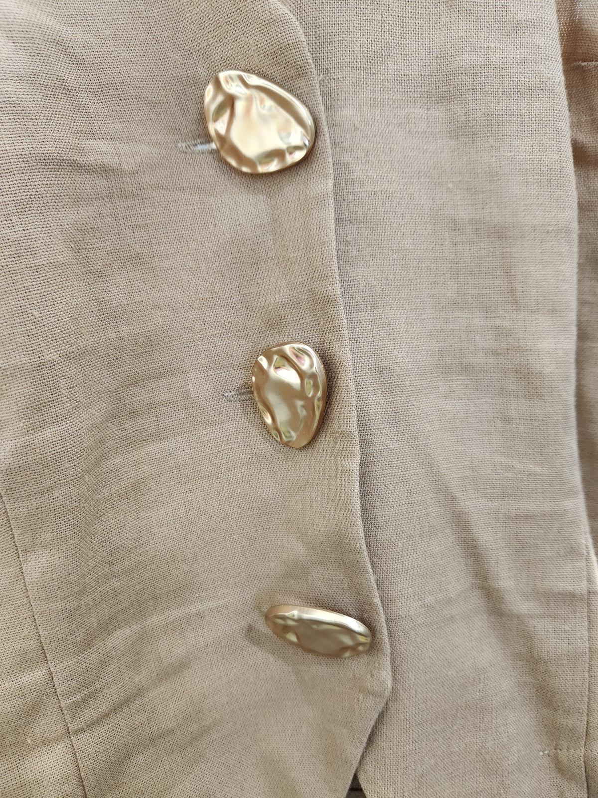 Close-up of a beige textured fabric garment with three large, organic-shaped metallic gold buttons.