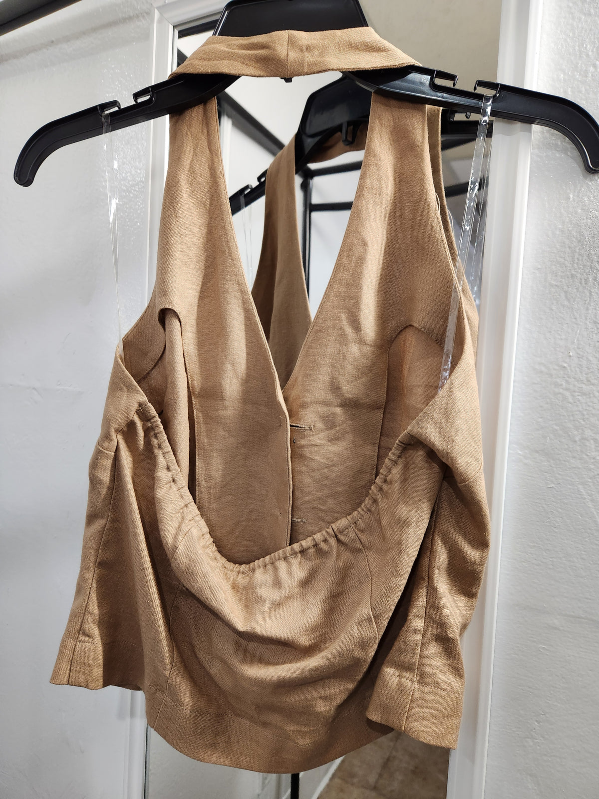 A tan linen halter top with a draped, gathered front design, hanging on a black hanger against a white door frame.