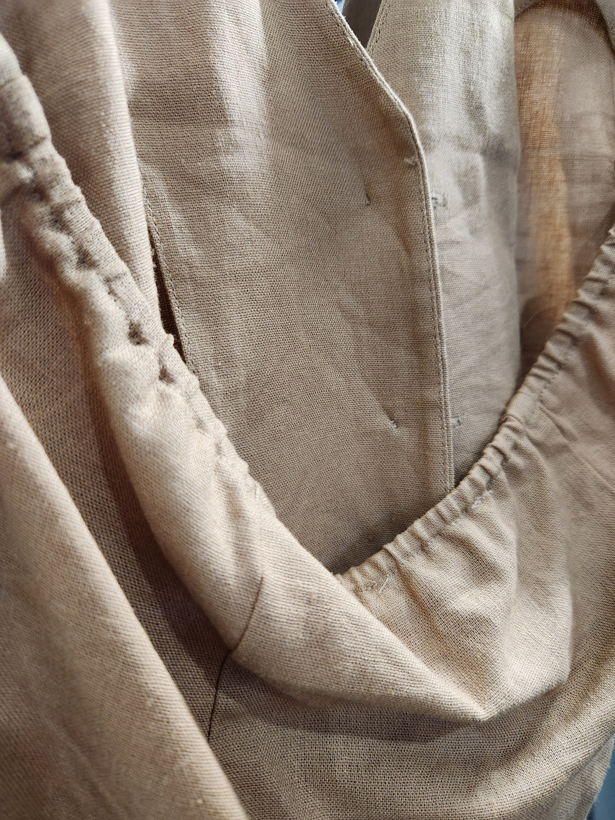 A close-up of a beige linen garment, showing the textured fabric, gathered elasticized edges, and a buttoned front closure.