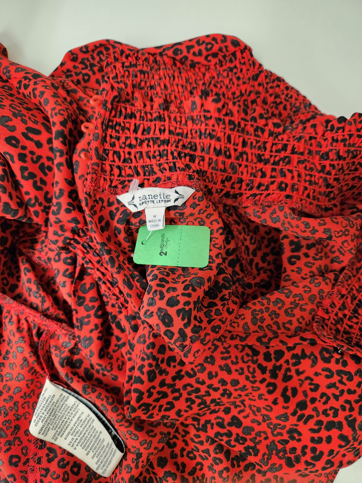 A close-up of a red and black leopard print garment, likely a dress or long top, featuring a smocked bodice and visible tags including a Janet Lee brand label and a green size tag.