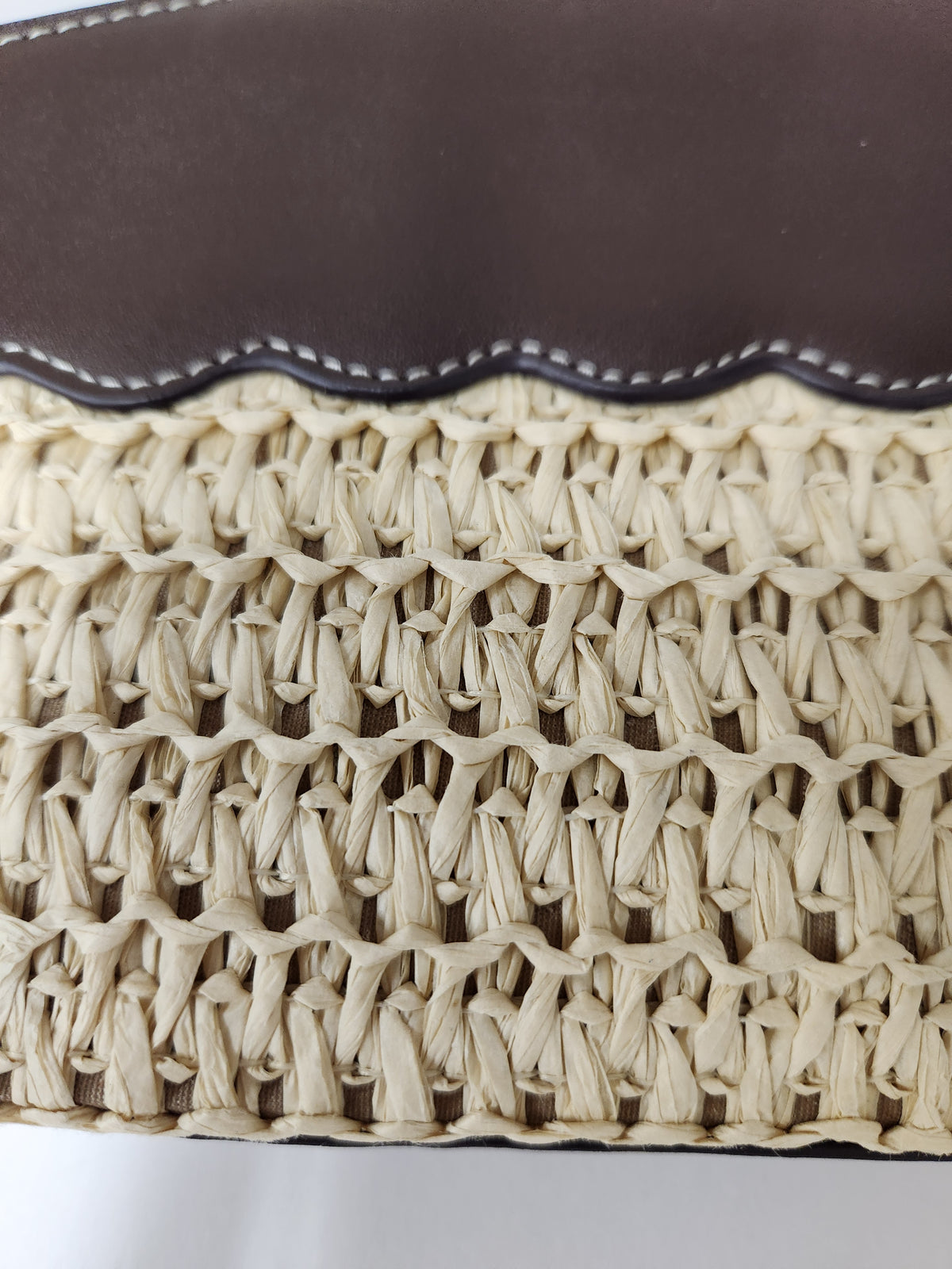 Close-up of a woven straw hat with a dark brown leather trim and white contrast stitching along the edge.