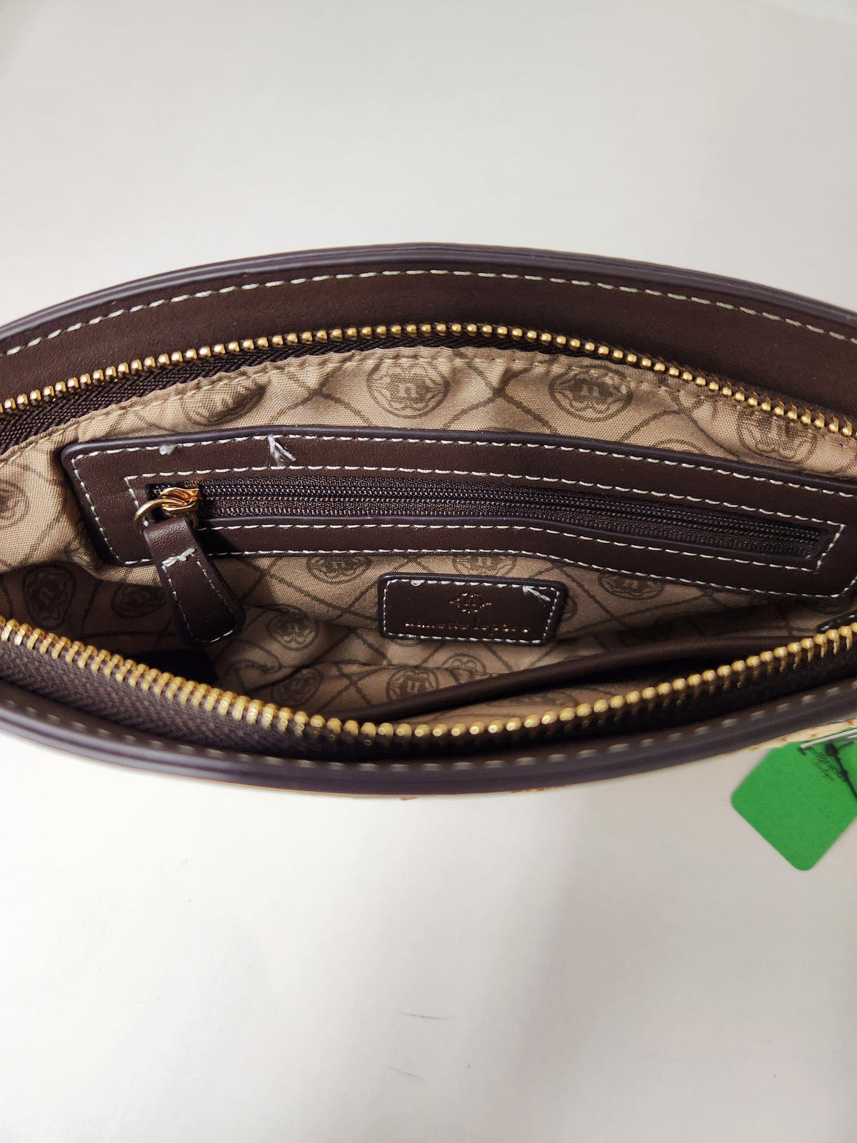 Close-up of the interior of a brown leather pouch or clutch, showing a patterned beige lining, a zippered pocket, gold-tone zipper teeth, and a small green tag attached near the edge.