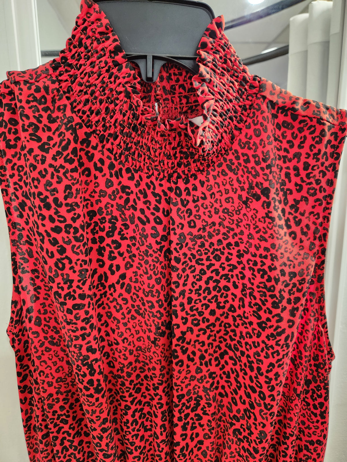 A sleeveless red top with a black leopard print pattern, featuring a smocked bodice and a high ruffled collar, shown hanging on a black hanger.