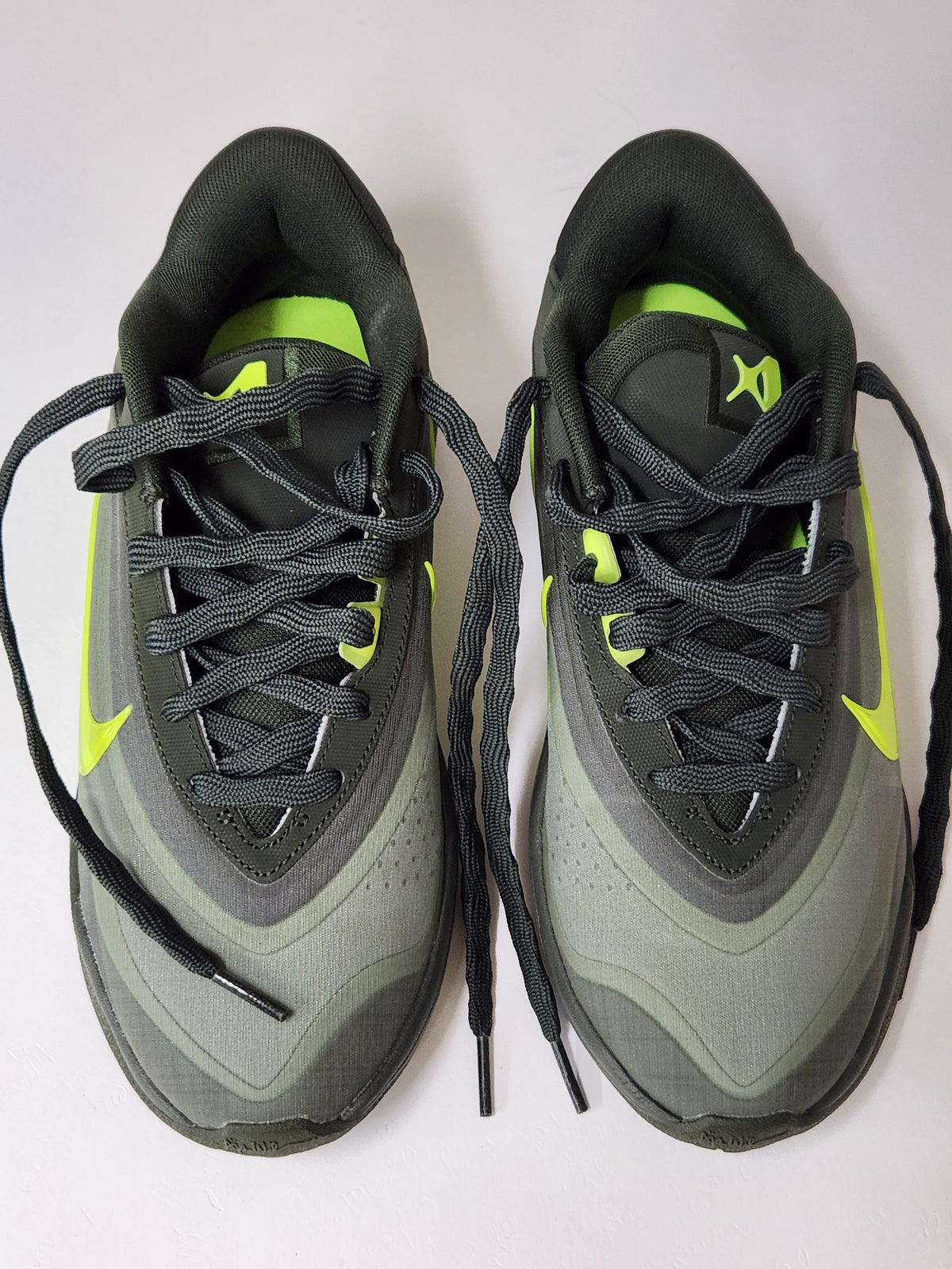 A pair of Nike low-top sneakers in dark gray and olive green with bright neon yellow-green accents, shown from a top-down view on a light background.