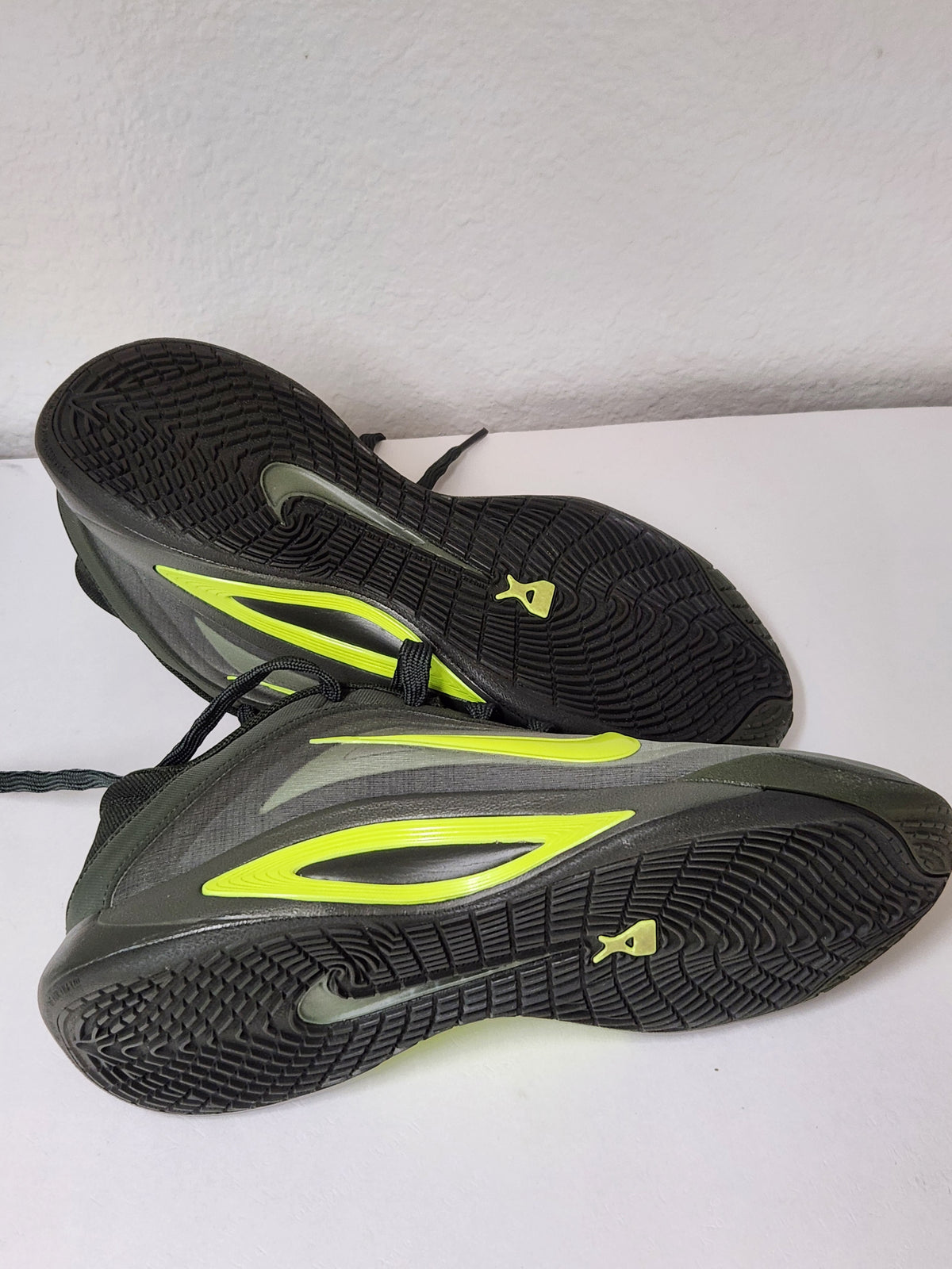 A pair of Nike running shoes shown from the bottom, featuring a black textured outsole with a small yellow Nike logo, a gray and black upper, and bright neon yellow-green accents.