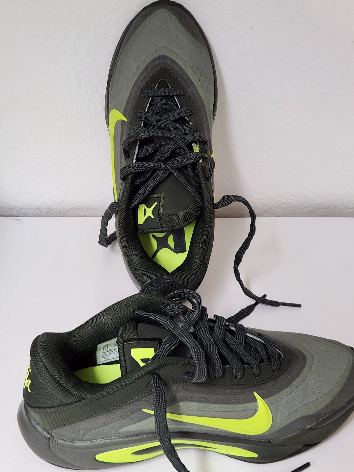 A pair of Nike running shoes in dark gray and black with bright neon yellow-green accents, displayed on a white surface against a white wall.