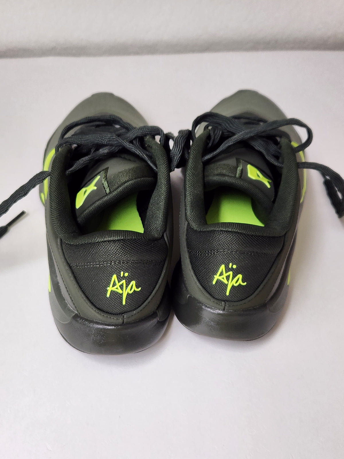 A pair of black low-top sneakers viewed from the back, featuring bright neon green accents including the 'Aja' script logo on the heel, a matching green insole, and green details on the tongue